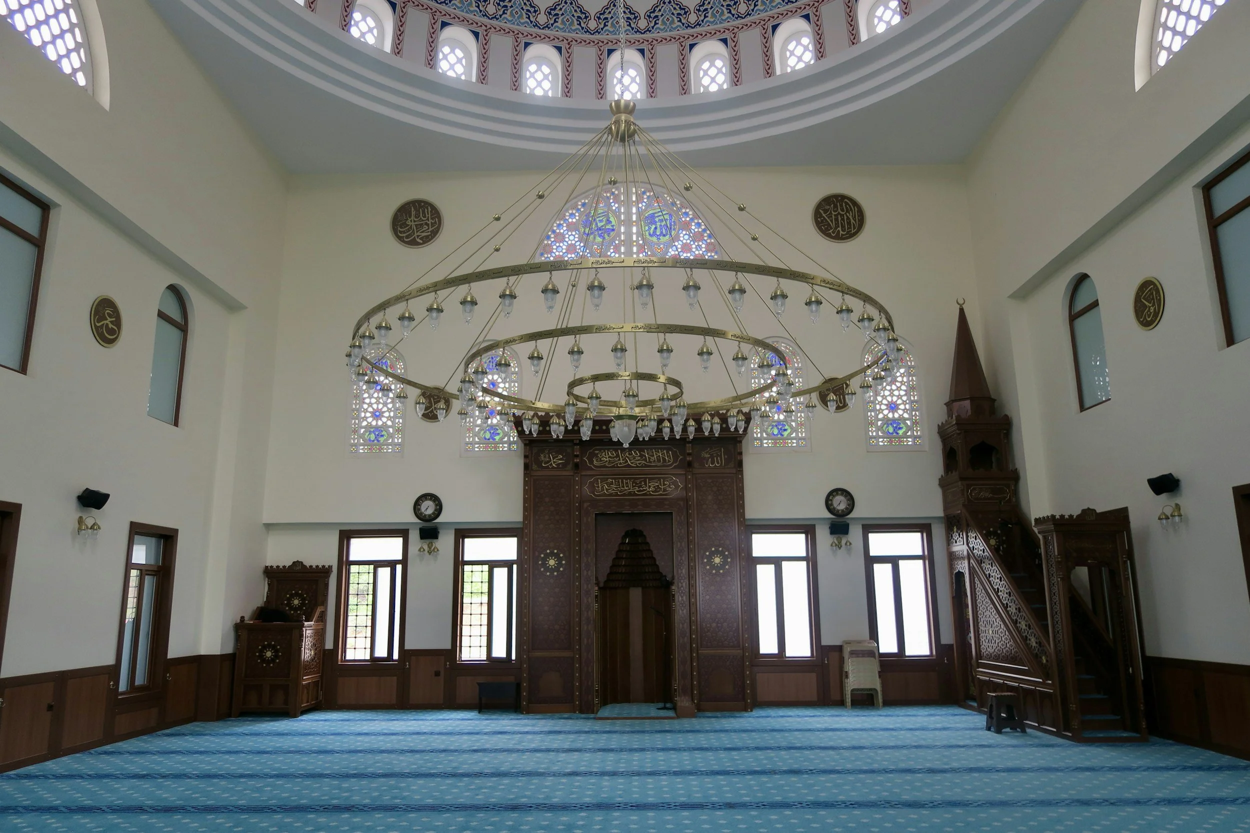 Interior of a mosque with a blue carpet, stained glass windows, and a large central chandelier hanging from the ceiling.