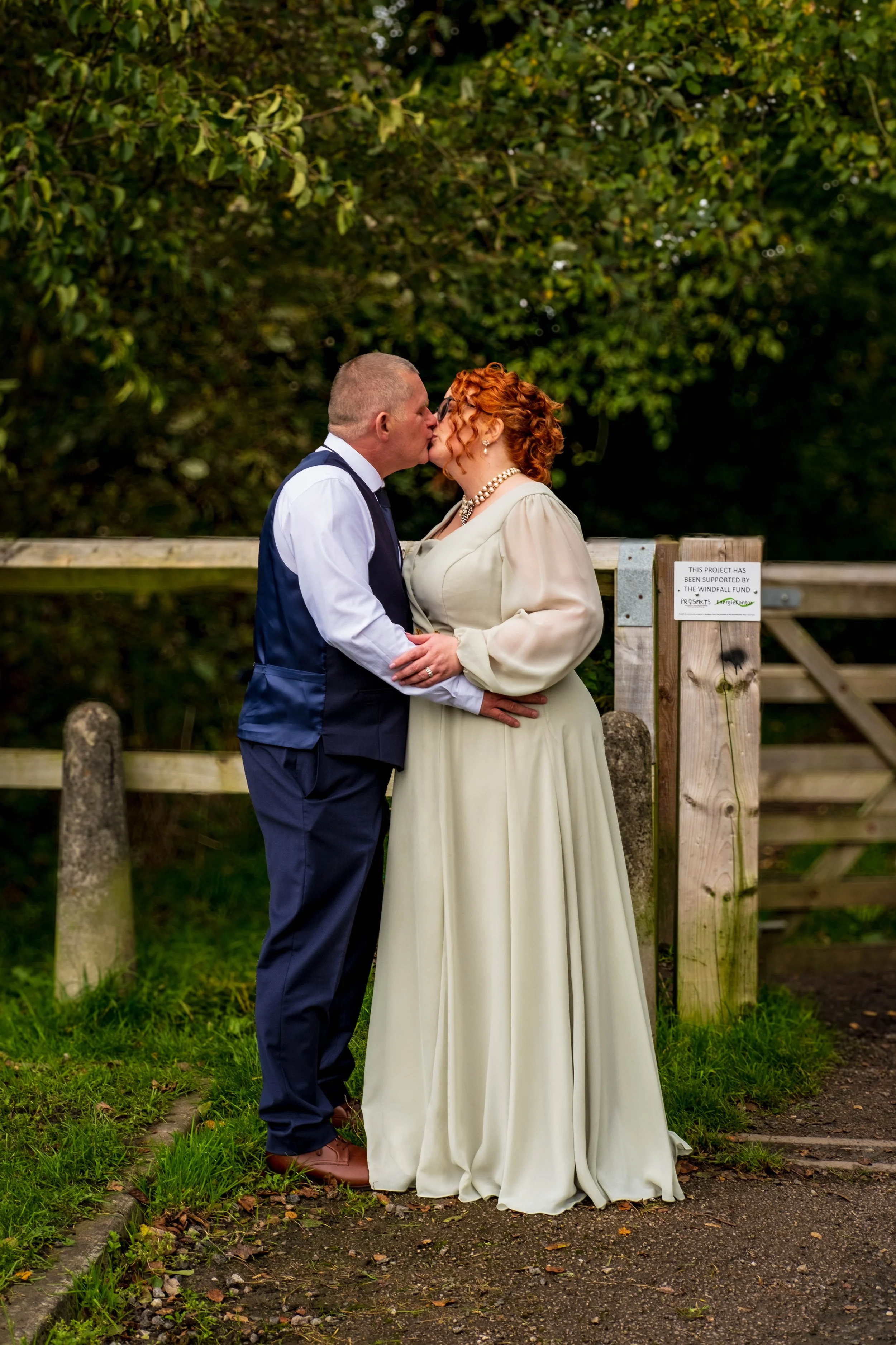 A couple kissing outdoors, dressed in wedding attire, standing on a grassy path near a wooden fence with green foliage in the background.