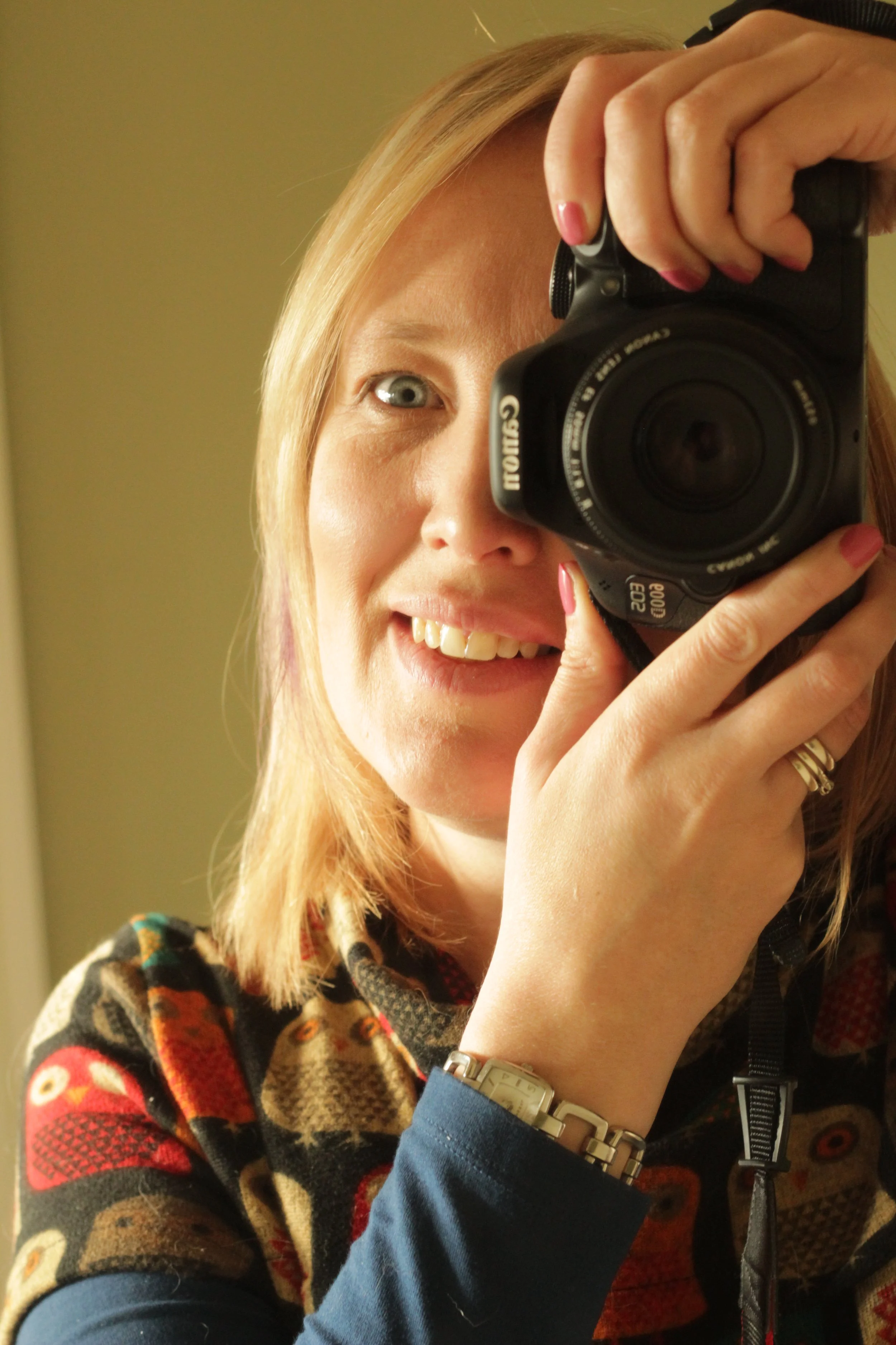 A woman with shoulder-length blonde hair taking a selfie using a Canon camera in front of a mirror, smiling and wearing a colorful patterned sweater.