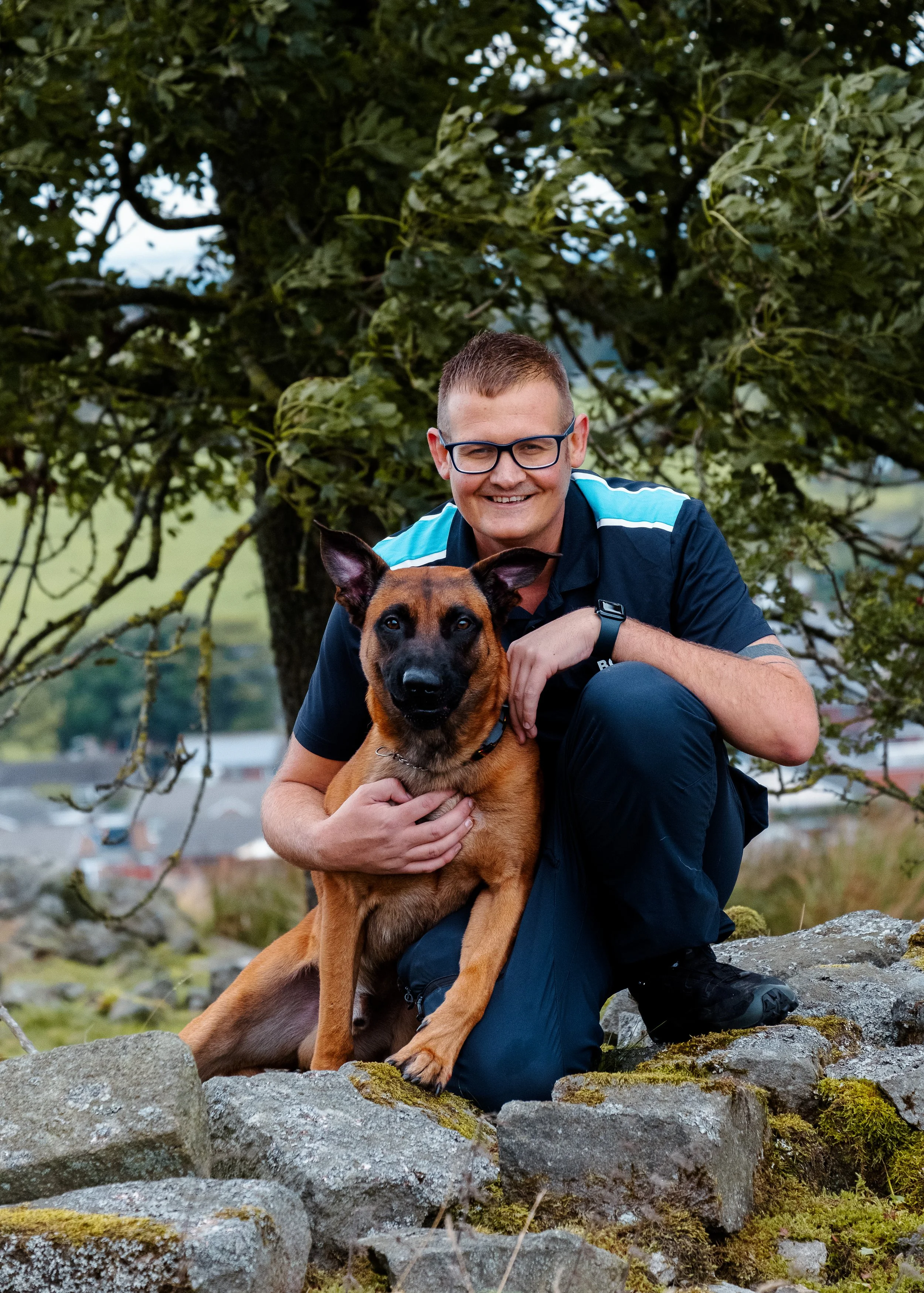A man with glasses and a short haircut kneeling on Rocks outdoors, smiling, with a large brown and black dog on a leash in front of him.