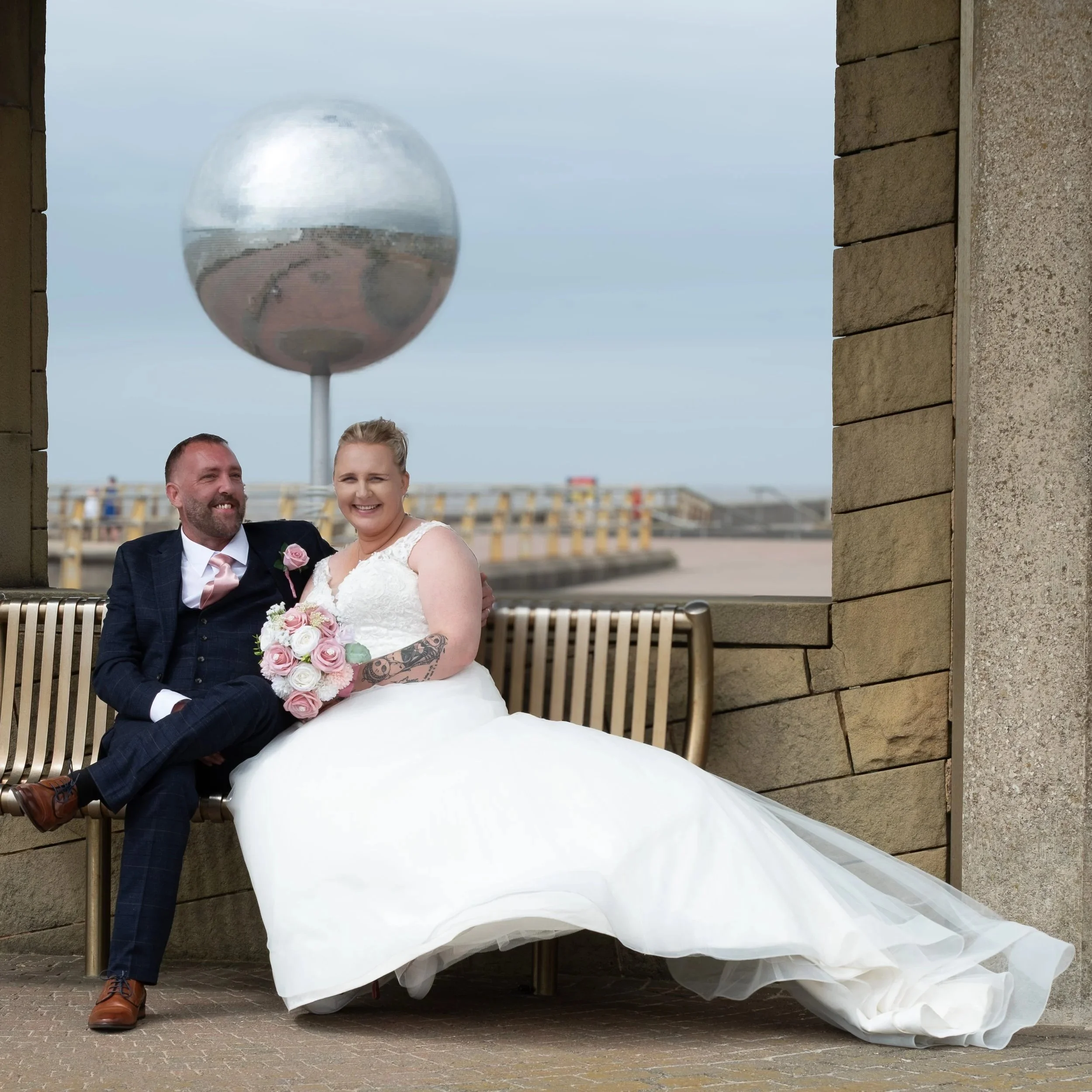 A newlywed couple sitting on a bench, smiling, with a large metallic spherical sculpture and an industrial waterfront in the background.