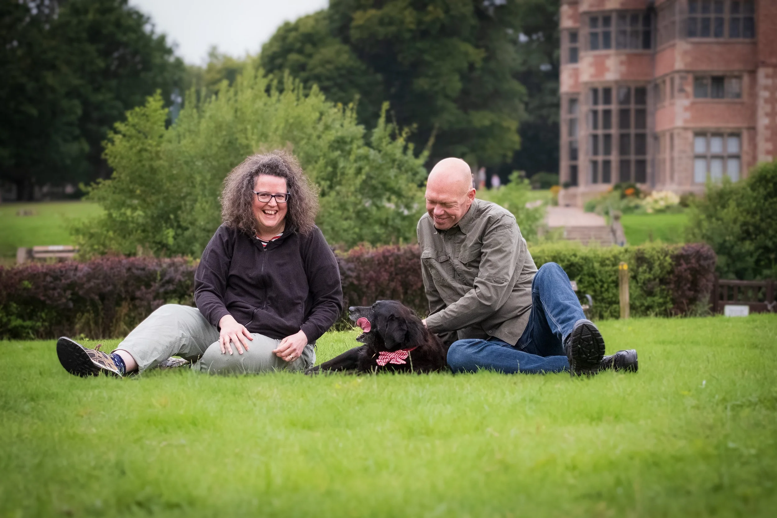 Two adults sitting on the grass and smiling with a black retriever dog, in a park with greenery and a large brick building in the background.