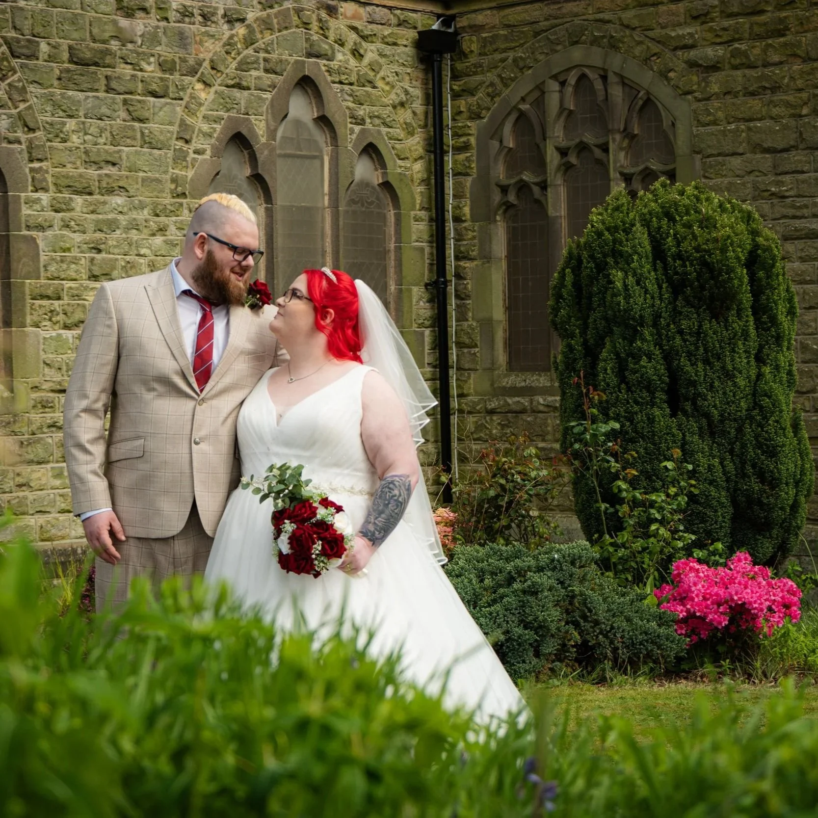 Bride and groom standing outside in front of an old stone building with arched windows, surrounded by greenery and pink flowers, sharing a moment at their wedding.