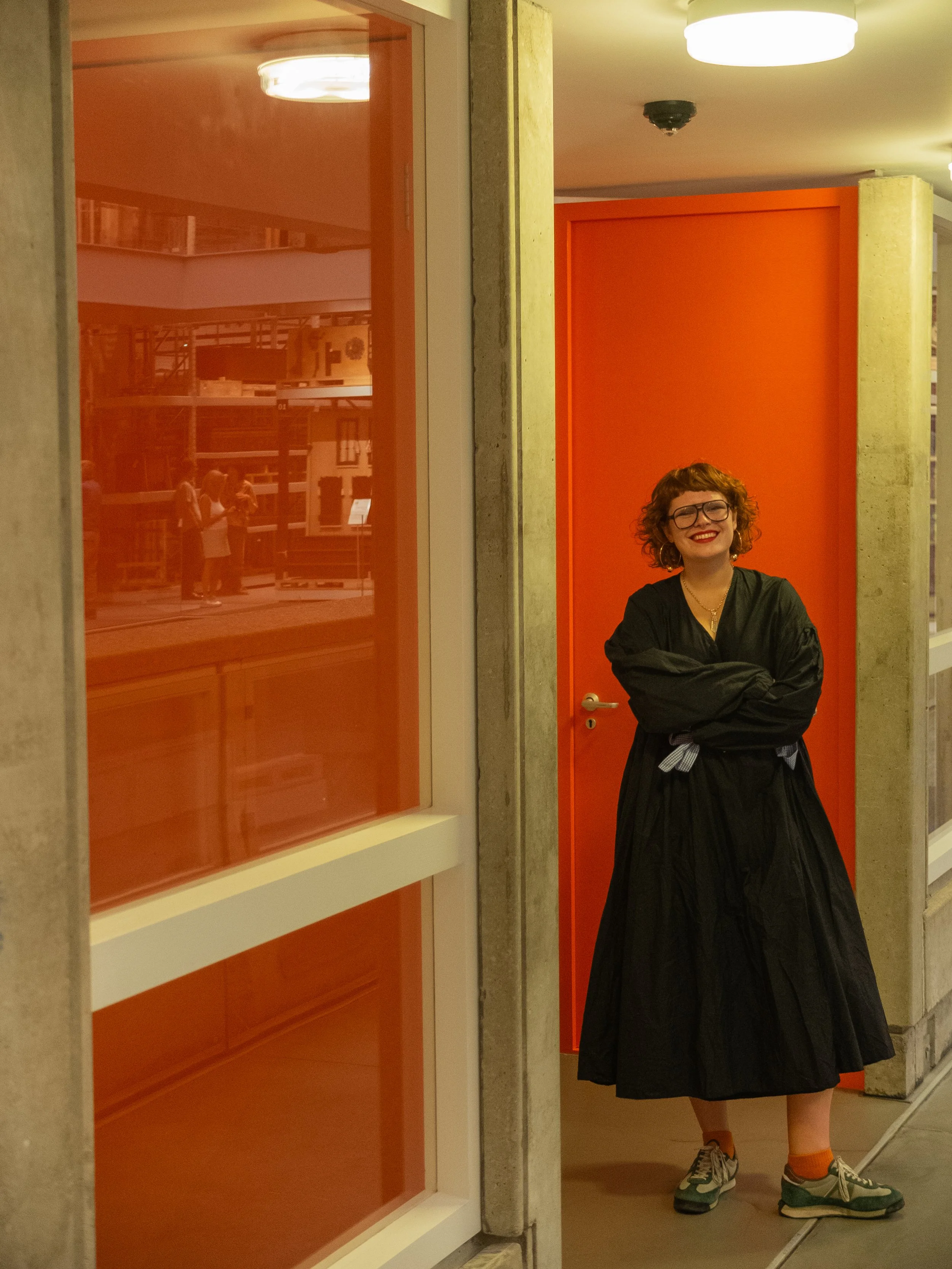 A woman with curly red hair, glasses, and a black dress is standing with arms crossed in front of an orange door in an indoor space, smiling.