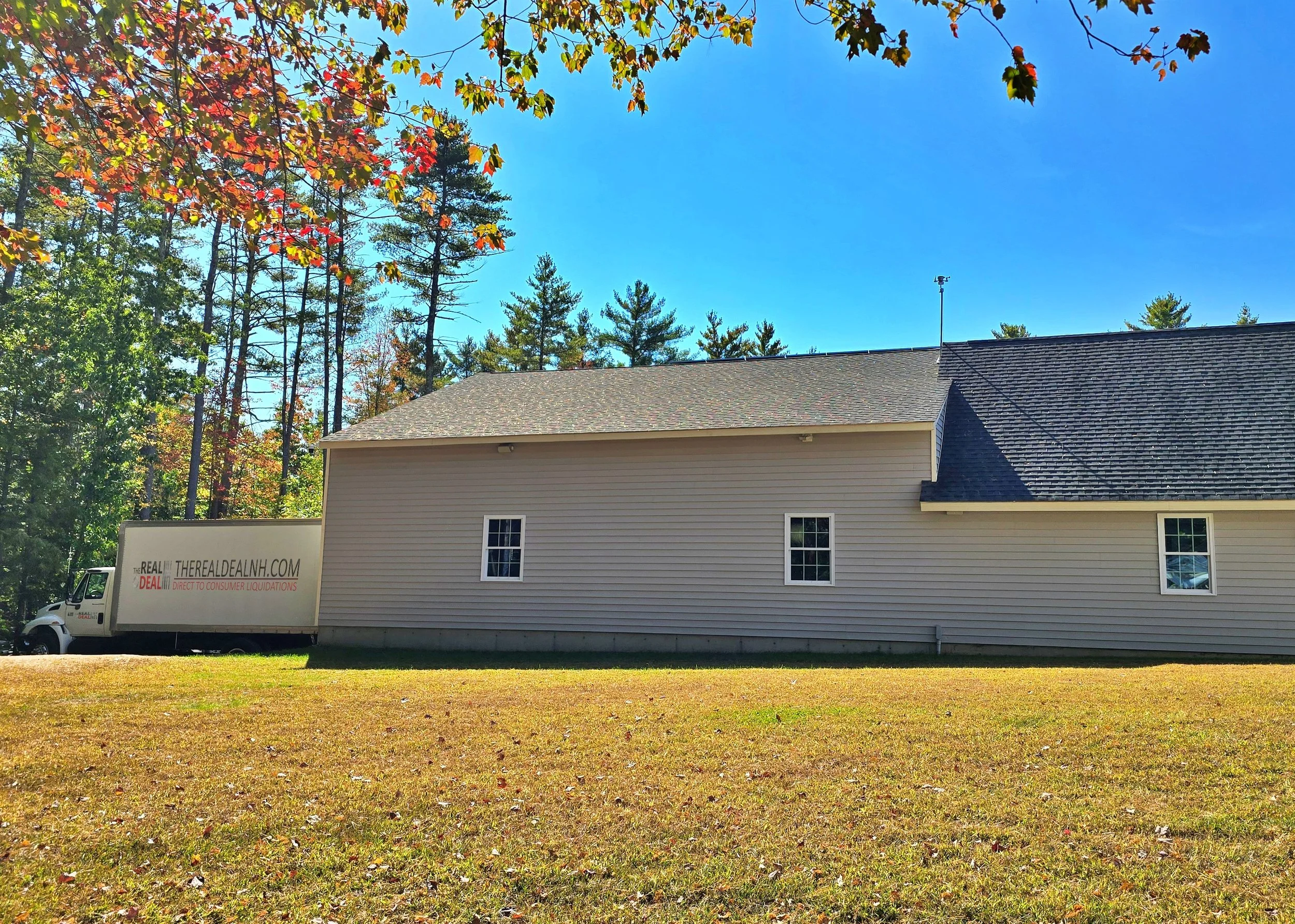 A beige house with a black roof, three windows on the side, a grassy yard, and a white truck with 'THEREALDEALNH.COM' written on it parked beside the house. Trees with fall foliage are in the background under a clear blue sky.