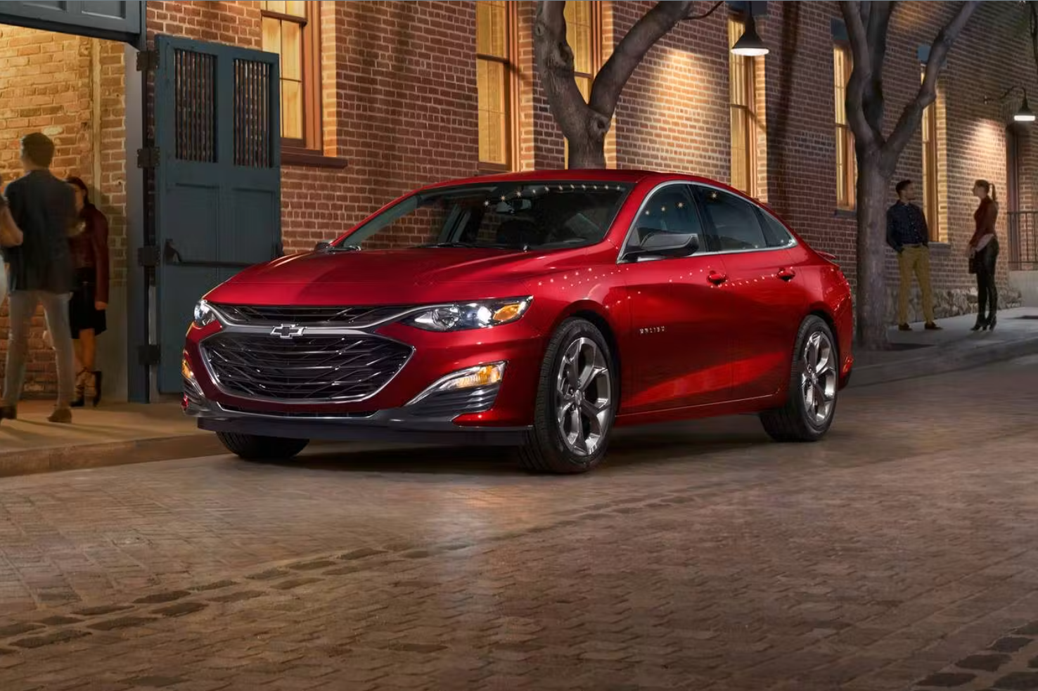 A red Chevrolet sedan parked on a city street at night with brick buildings, trees, and a few people standing nearby.