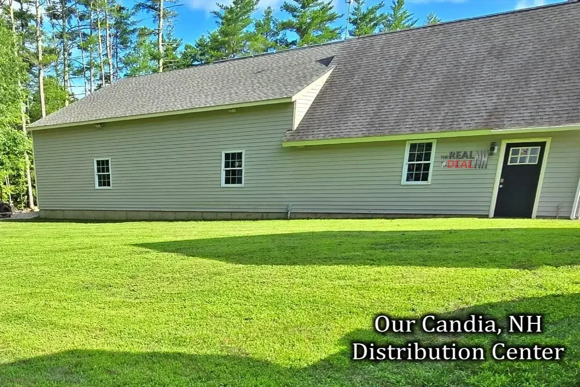 A beige building with three small windows and a black door, located in a grassy yard with a forest of trees in the background. Text in the bottom right corner says, "Our Candia, NH Distribution Center."