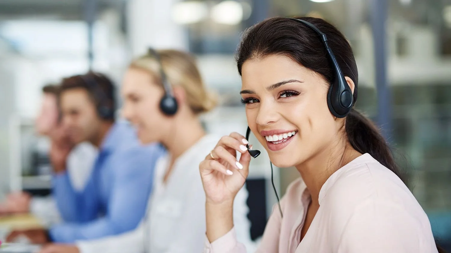 A woman smiling while wearing a black headset, with three other colleagues in the background also wearing headsets, in a bright office setting.