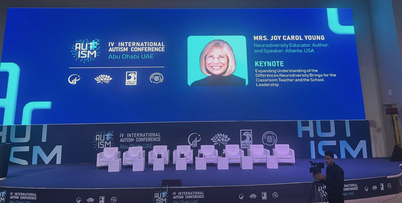 Stage and giant screen at 2026 IV International Autism Conference, showing Joy Carol Young profile photo in a black turtleneck and with a title of Keynote.