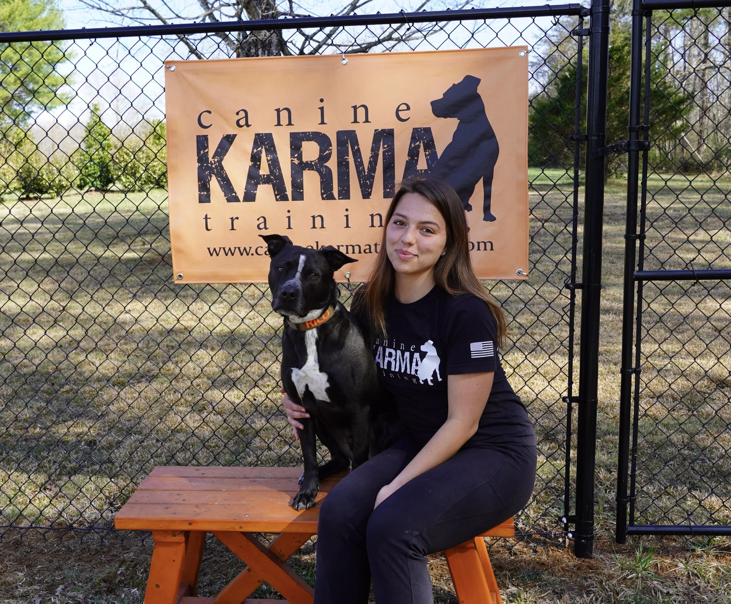 A woman sitting on an orange picnic table holding a black and white dog, in front of a fence with a sign that reads 'canine KARMA training' and features a silhouette of a dog. The background shows trees and grass.