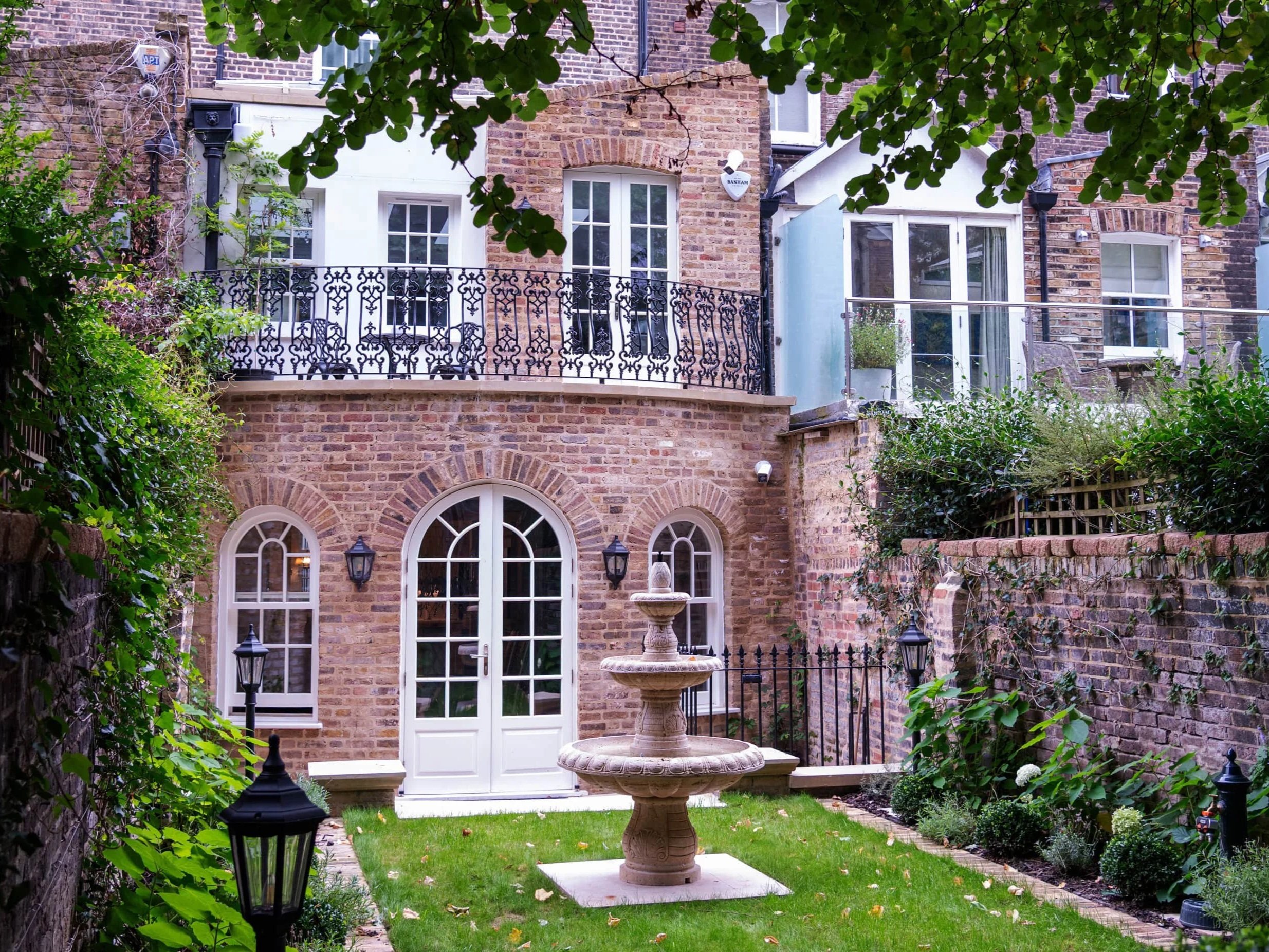 Backyard garden with a fountain, surrounded by greenery, brick walls, and a white house with a balcony and large windows.