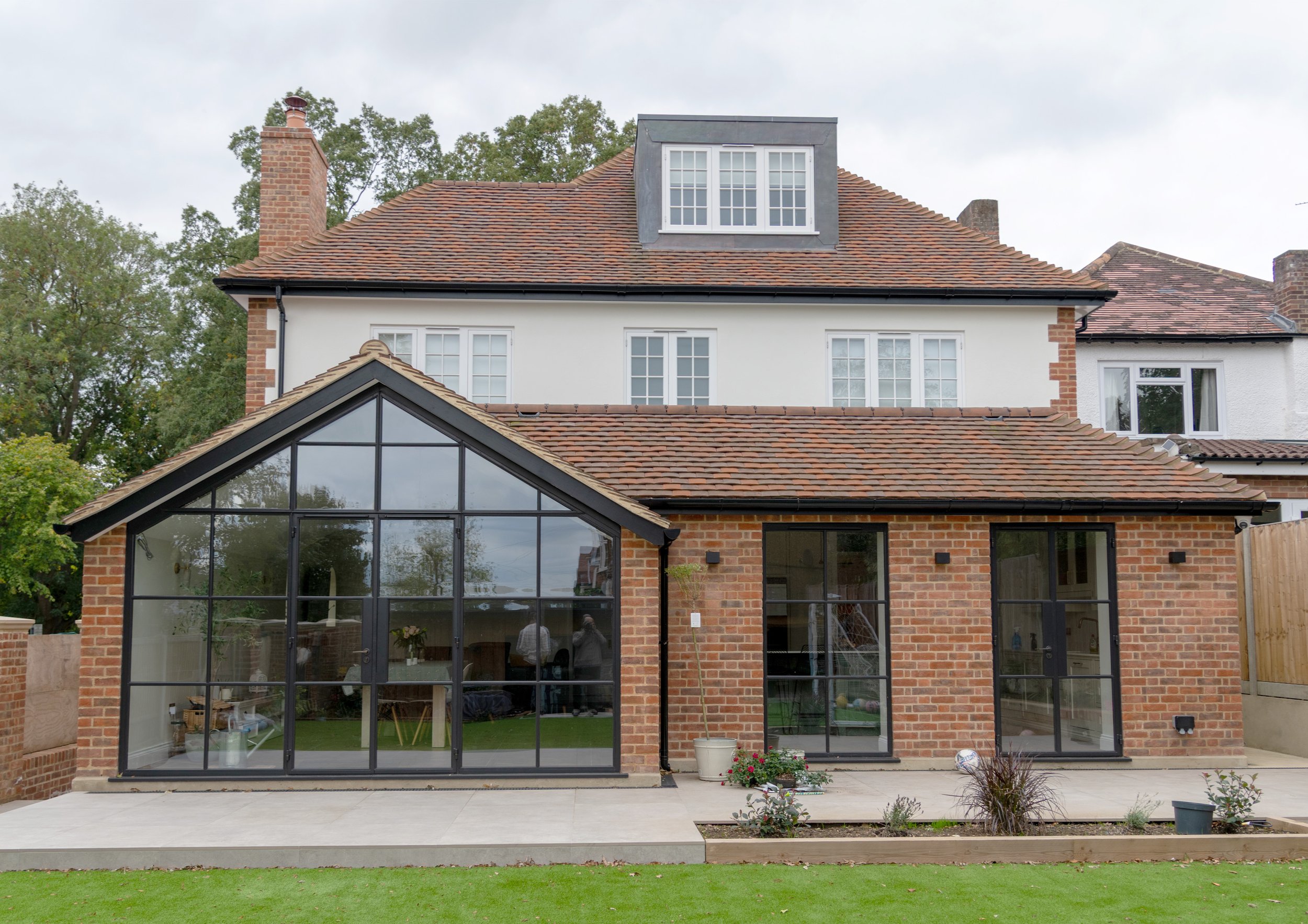 The back of a house with a brick exterior and large glass windowed conservatory, a backyard garden, and trees in the background.