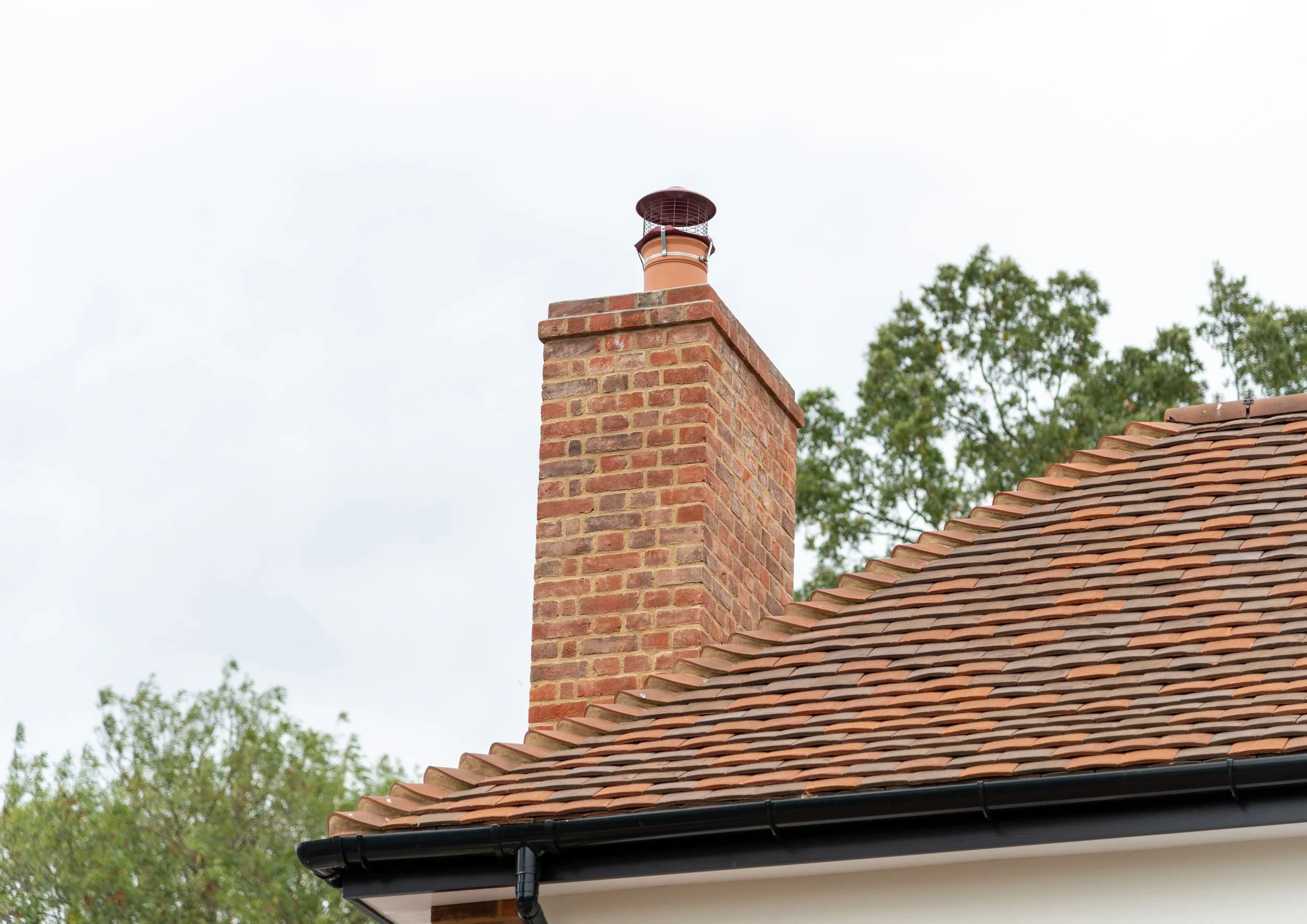 Close-up of a brick chimney with a cap on top, attached to a house with a sloped terracotta tiled roof and a black gutter.