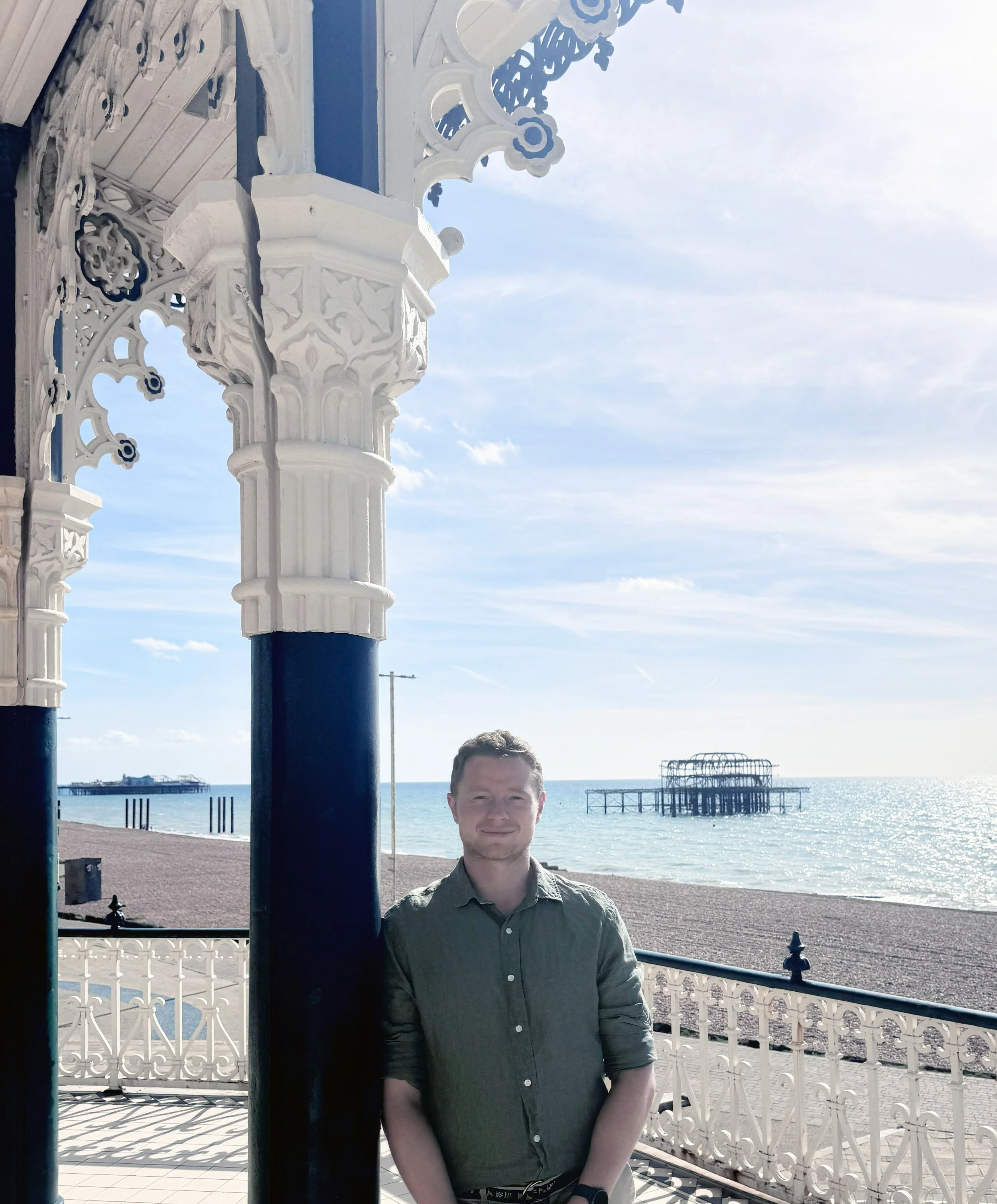 A man standing on a Victorian-style balcony with intricate white wrought iron railings, overlooking a beach and ocean, with a pier in the distance under a partly cloudy sky.