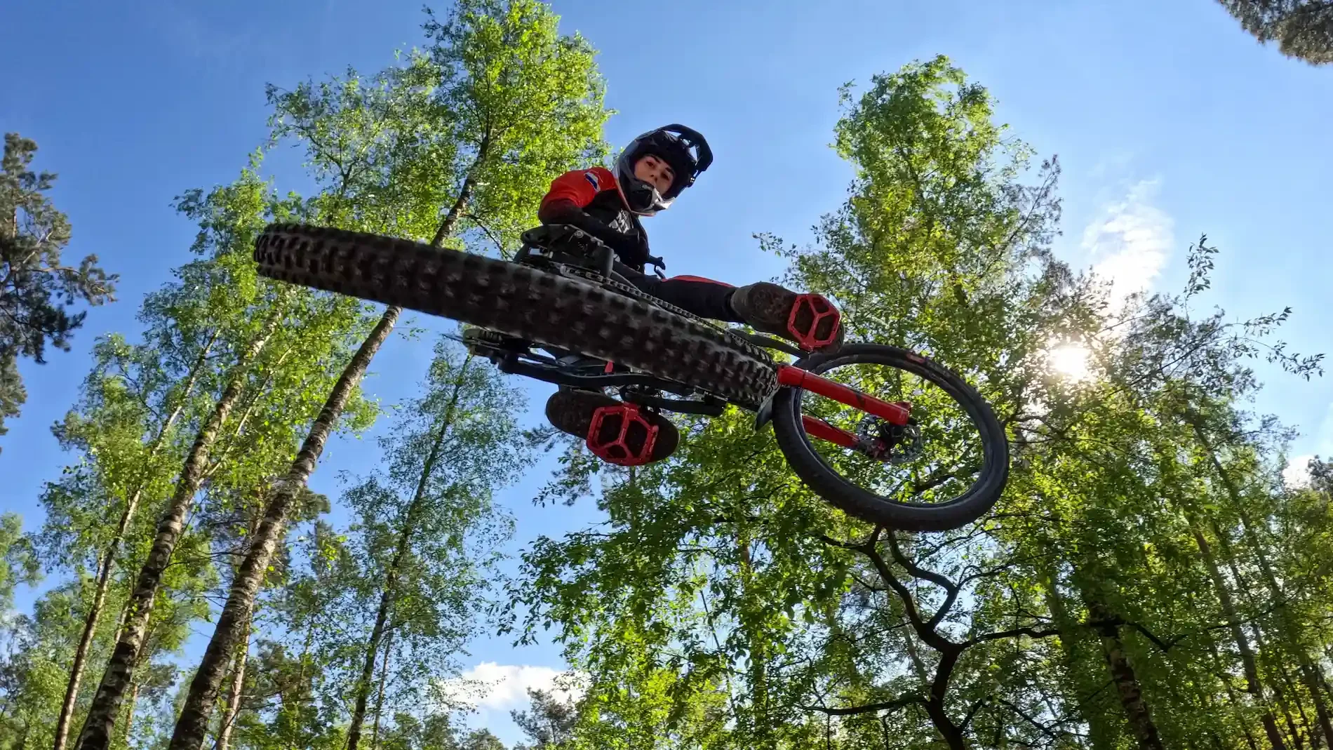 Deel van een mountainbike in de lucht tussen groene bomen en een blauwe lucht, met een jonge man in een helm die op de mountainbike rijdt.