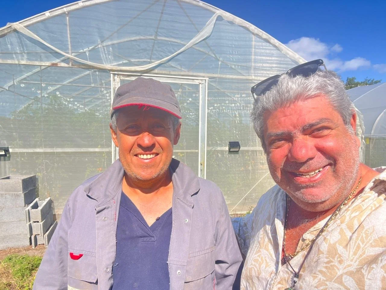 Deux hommes souriants prennent un selfie devant une serre en plastique sous un ciel bleu, l'un porte un chapeau gris avec une visière rouge, l'autre des lunettes de soleil sur la tête.