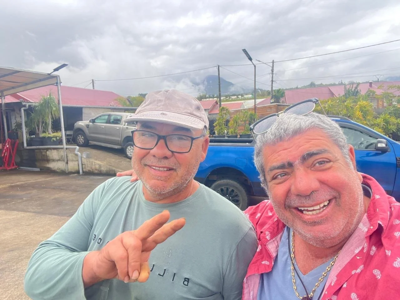 Deux hommes souriants posant pour une photo en extérieur avec des véhicules et des maisons en arrière-plan, un ciel nuageux.