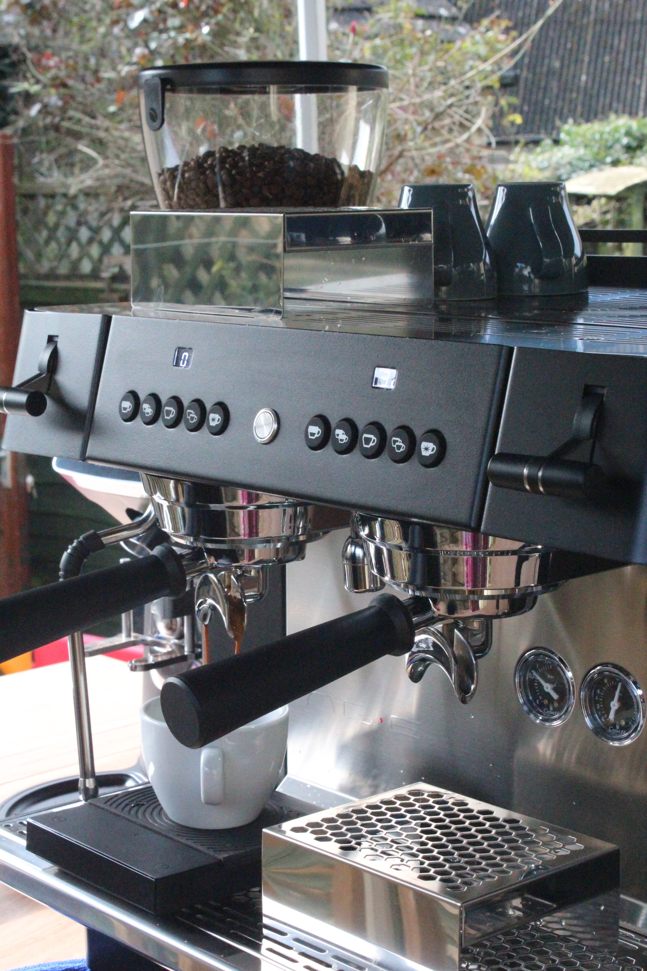 A professional espresso machine with multiple brew heads, a portafilter dispensing coffee, and a cup underneath collects the espresso, set outdoors with a window and garden in the background.