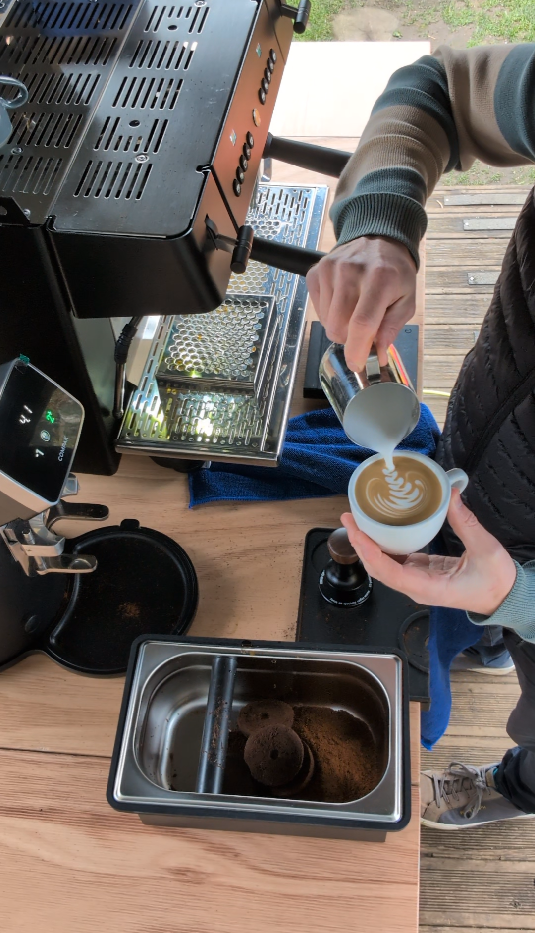 A person pouring steamed milk into a cup of espresso, creating latte art, on a wooden table outside. Breweries, coffee beans, and a scene of outdoor flooring are visible.