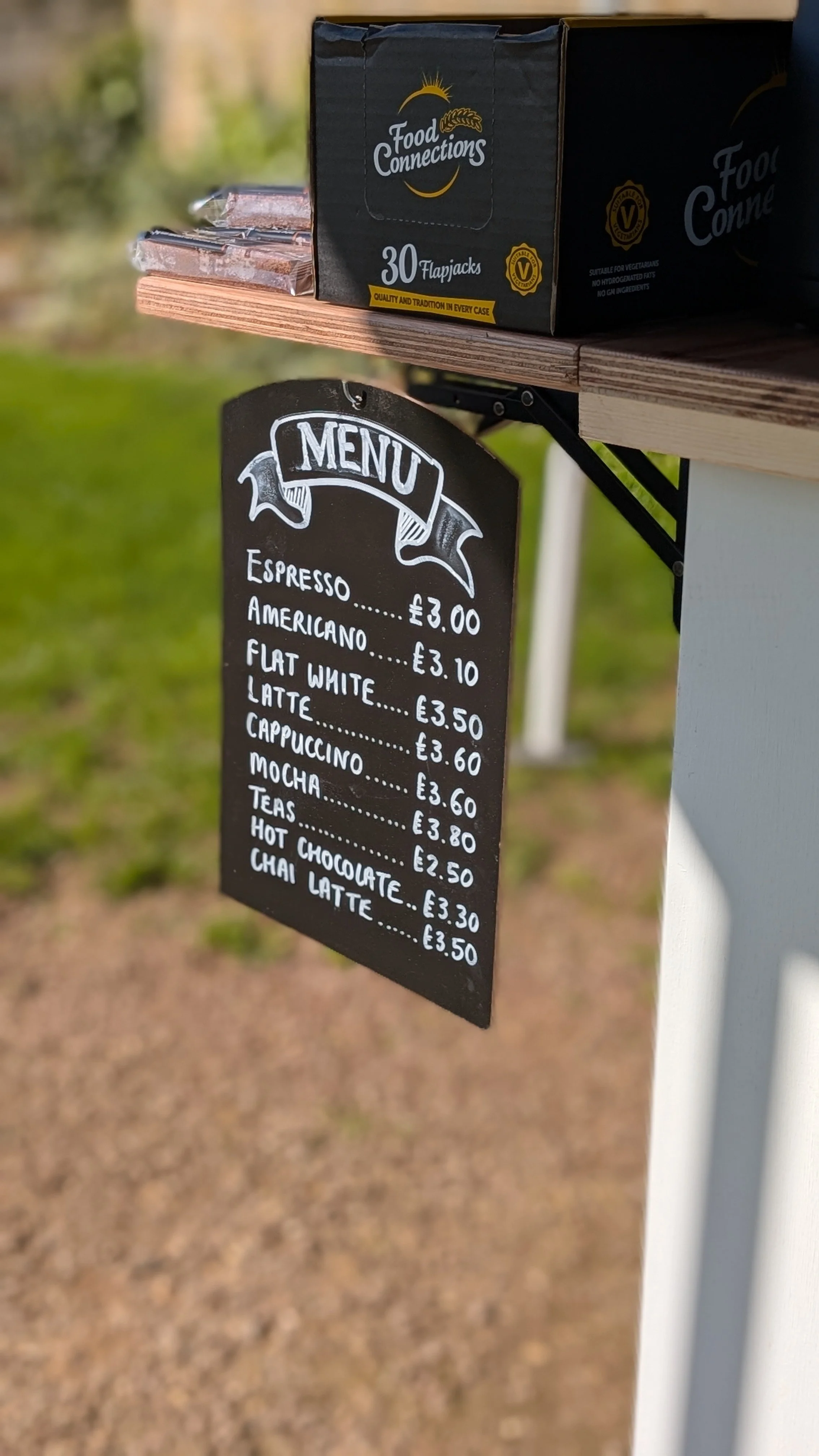 A chalkboard sign with a menu of coffee and tea drinks, with prices, hanging from a wooden stand outdoors. The menu includes espresso, Americano, flat white, latte, cappuccino, mocha, teas, hot chocolate, and chai latte.