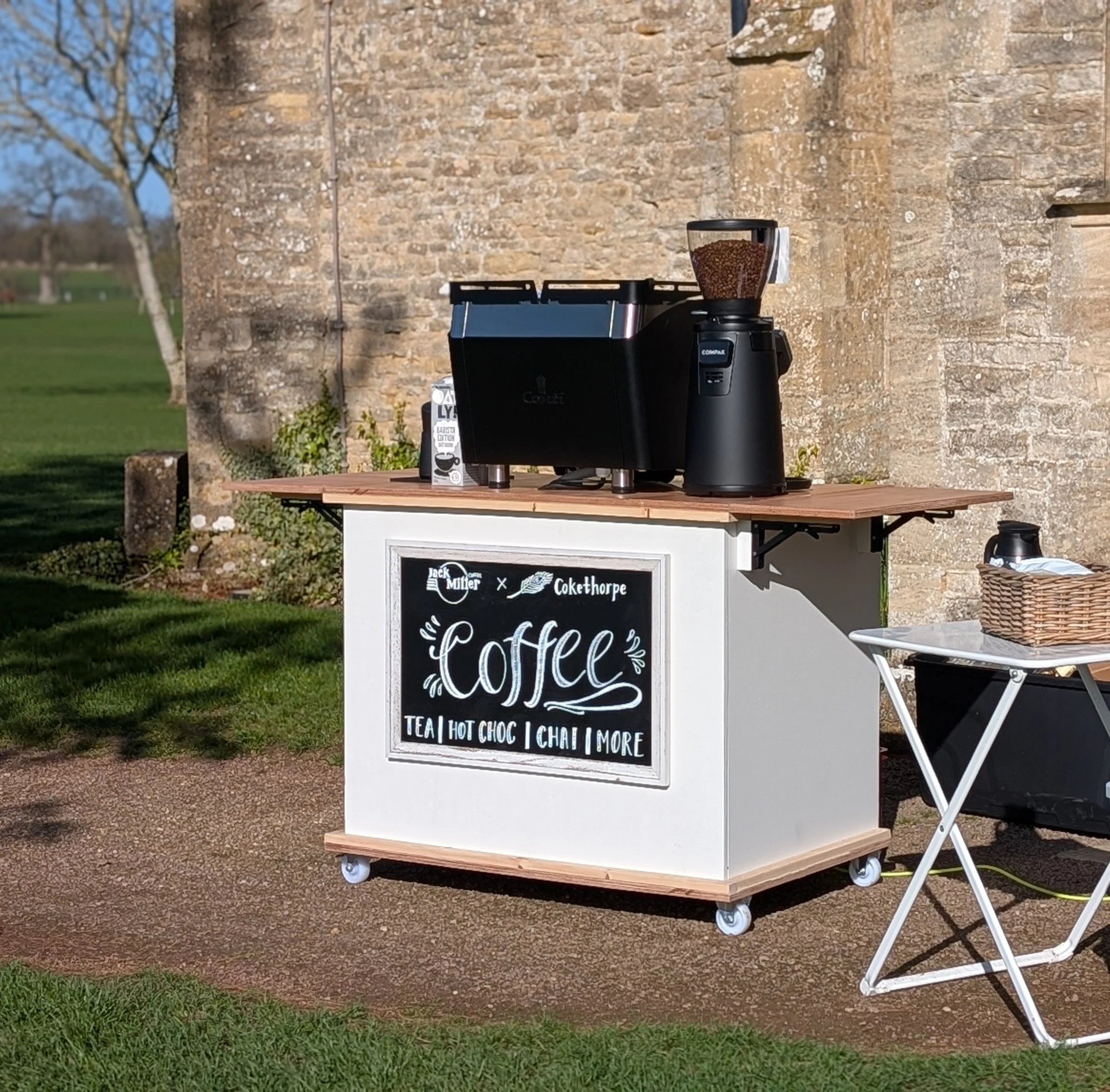 A portable coffee stand with a black signboard displaying coffee offerings such as tea, hot chocolate, chai, and more, set against an outdoor backdrop with grass and a stone wall.