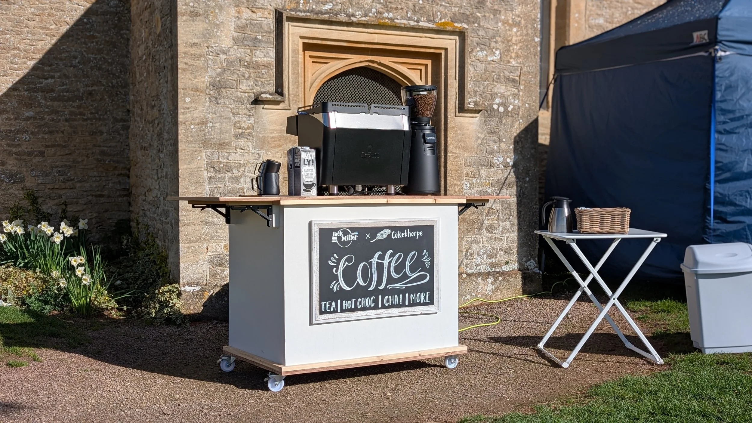 Coffee stand with coffee machine and supplies, chalkboard sign advertising tea, hot chocolate, chai, and more, in front of stone building, flowers nearby, outdoor setting.