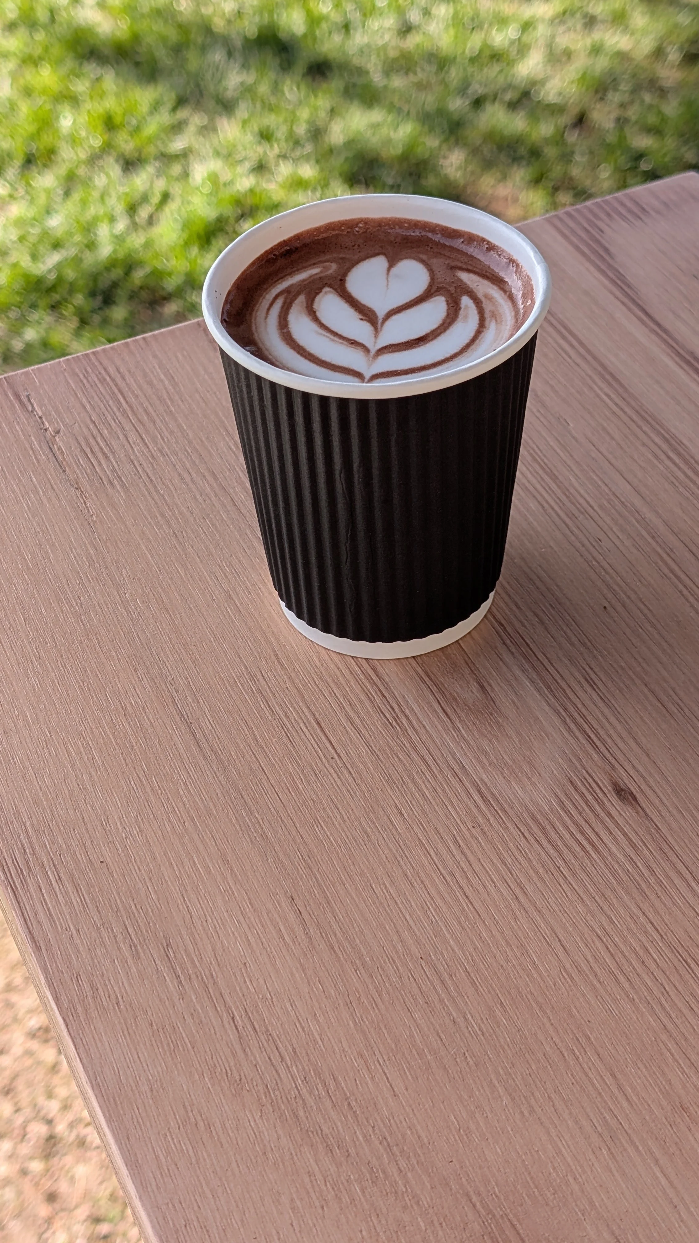A paper cup of hot chocolate with a latte art heart and leaf design, placed on a wooden table outdoors with green grass in the background.