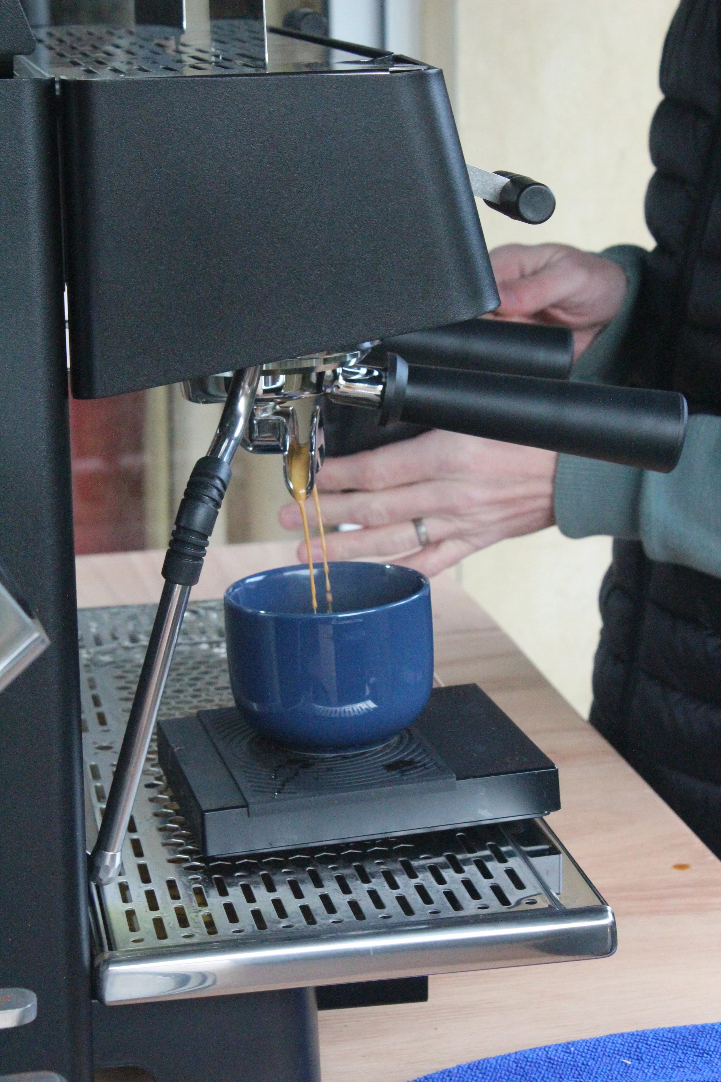 A person brews espresso coffee using a machine, with espresso flowing into a blue cup.