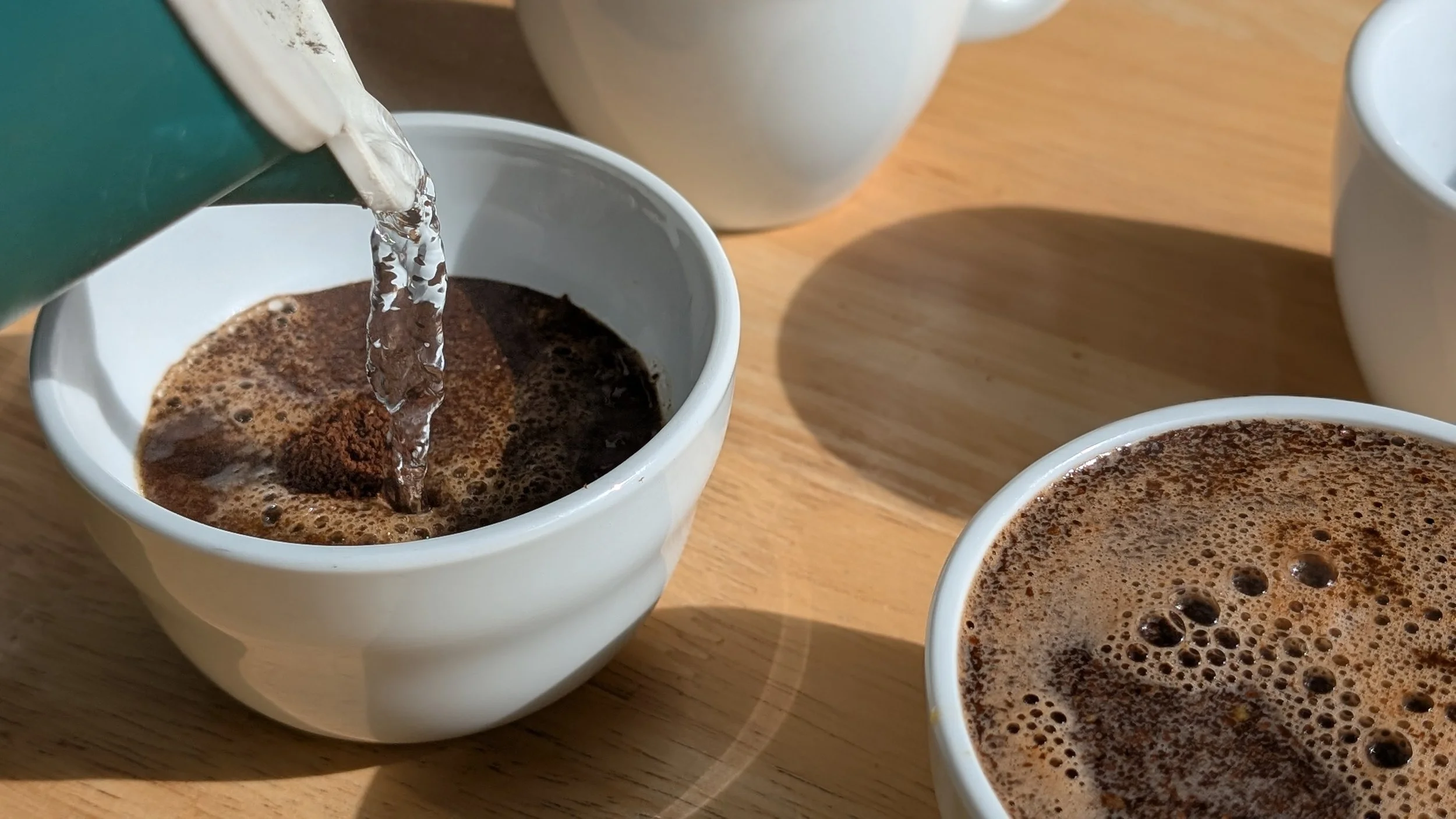 Hot water being poured into a cupping bowl with ground coffee on a wooden table with additional cups of coffee nearby.