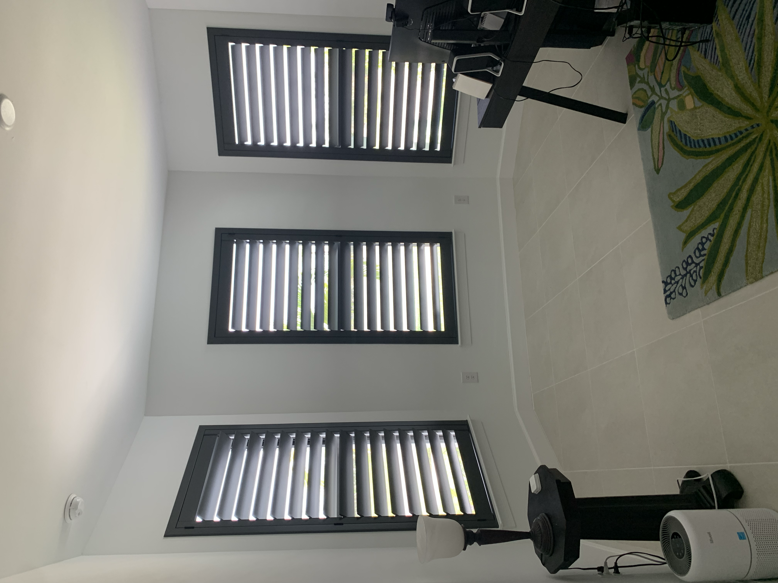 Empty room with three windows with black plantation shutters, a black desk with computer monitor and speakers, colorful rug with leaf pattern, and a white air purifier.
