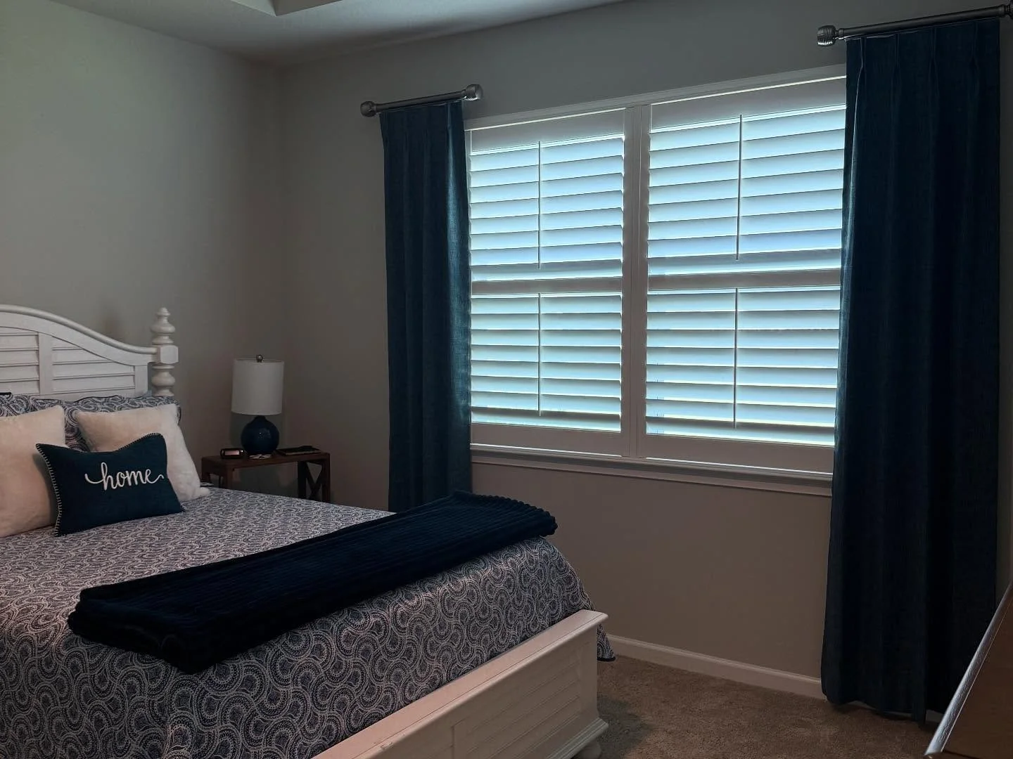 Bedroom with a white bed frame, patterned bedding, a navy pillow with the word 'home,' a side table with a blue lamp, and closed white plantation shutters with dark blue curtains.