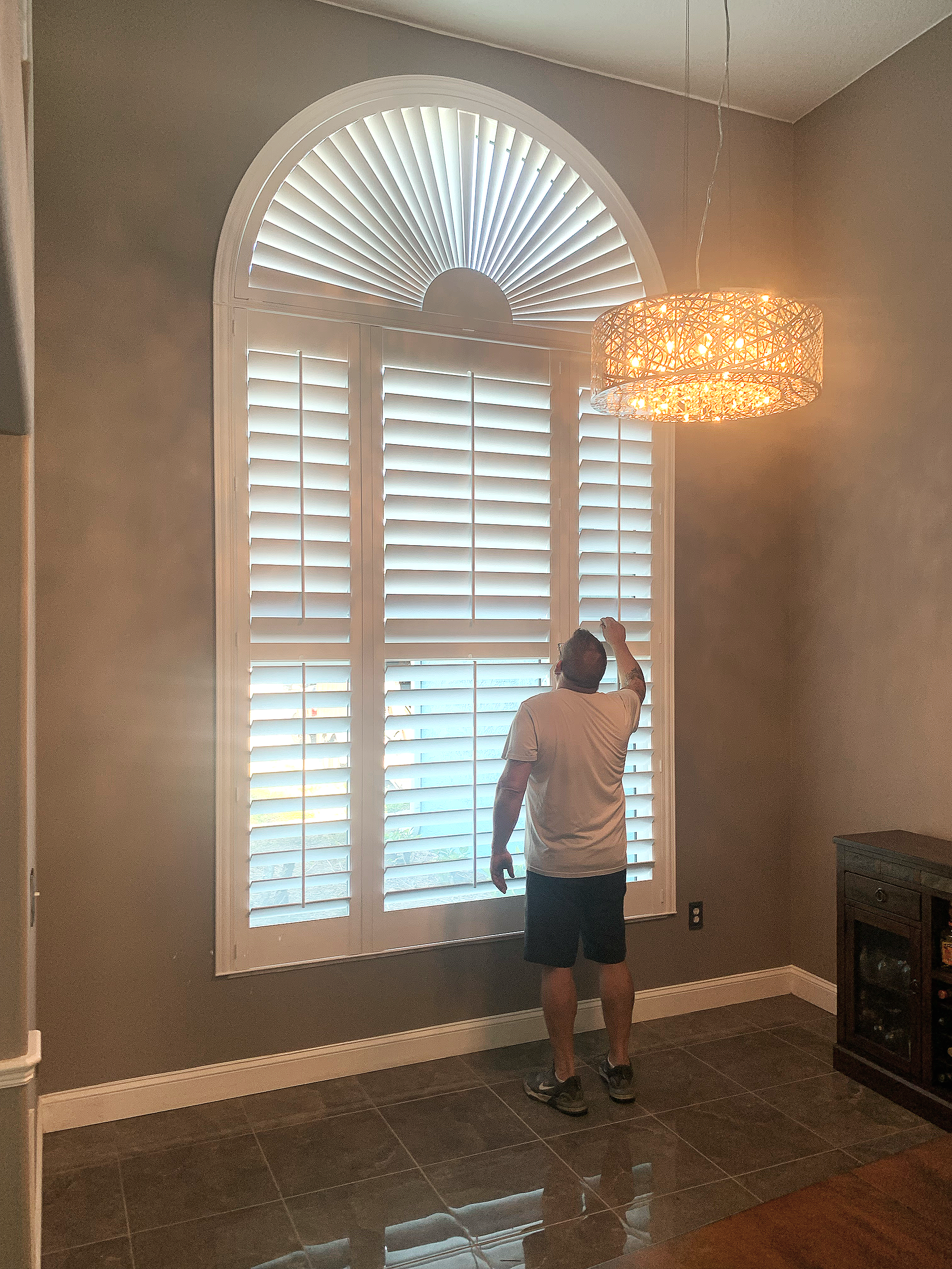 Man adjusting plantation shutters on a large window with an arched top, in a living room with tiled dark flooring and a hanging light fixture.