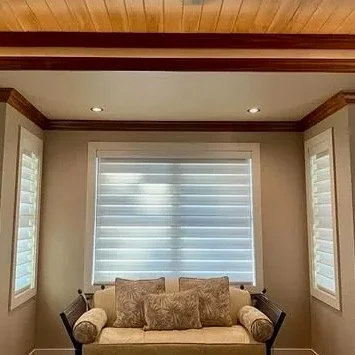 A living room with beige walls, large white window with shades, and a beige sofa with decorative pillows.