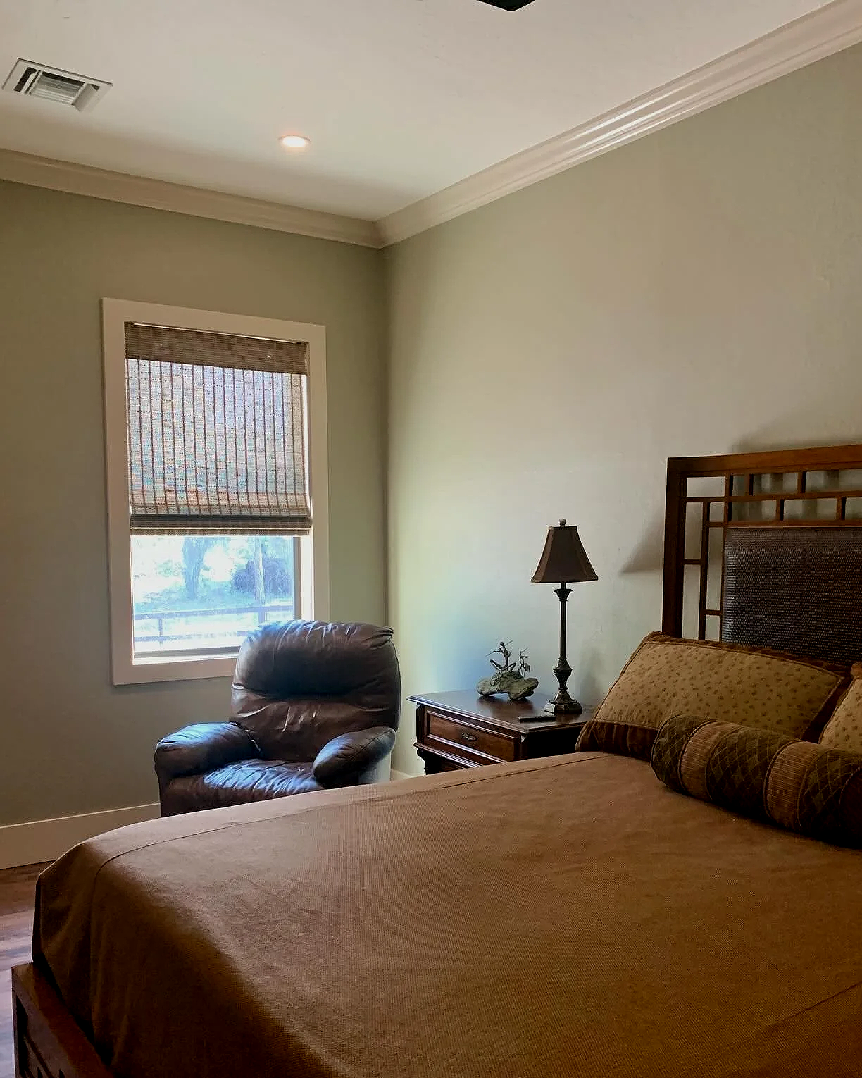 A bedroom with a bed, nightstand, lamp, leather chair, a window with a woven blind, and wood flooring.