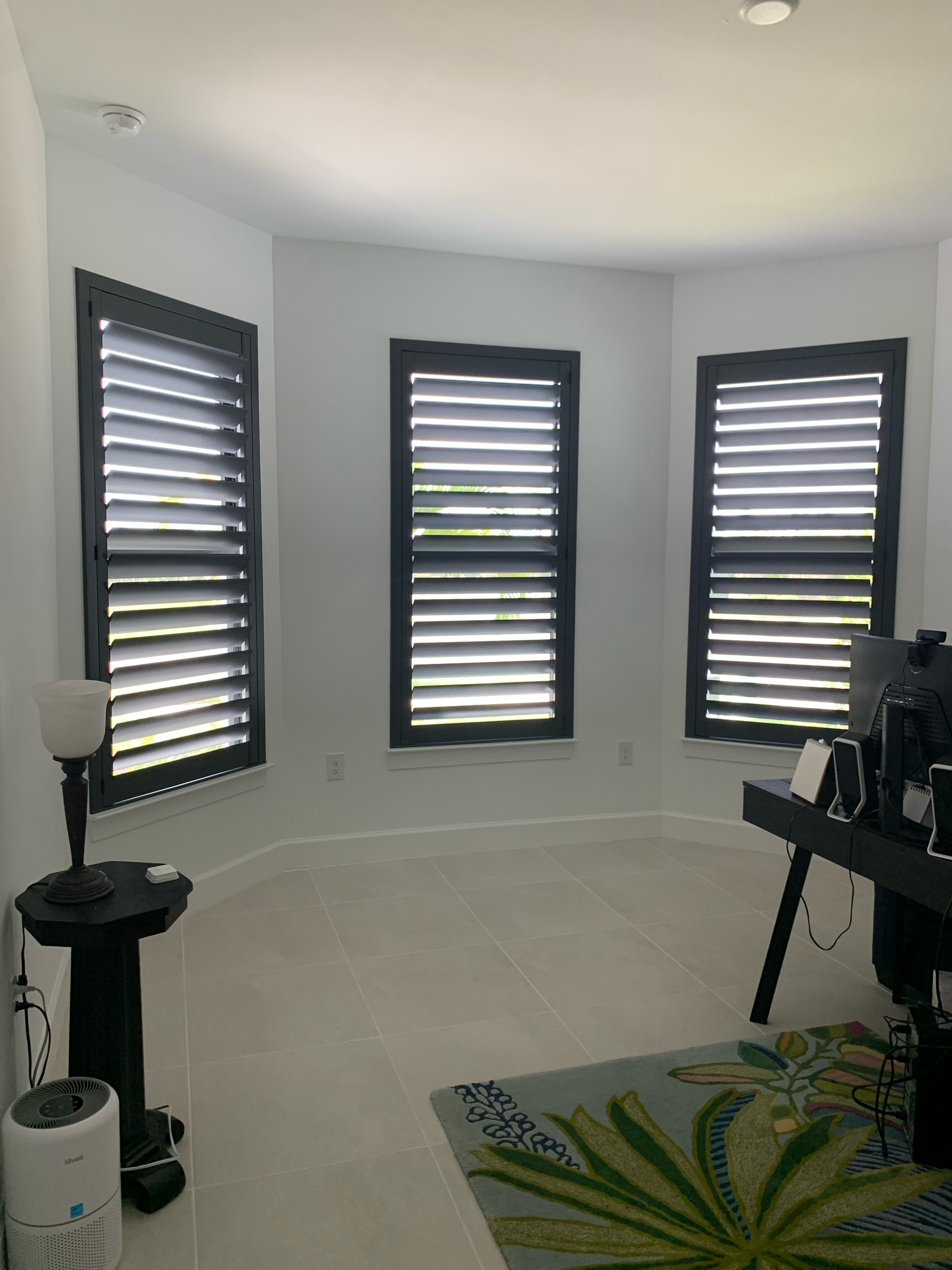 Empty room with three windows with black plantation shutters, a black desk with computer monitor and speakers, colorful rug with leaf pattern, and a white air purifier.