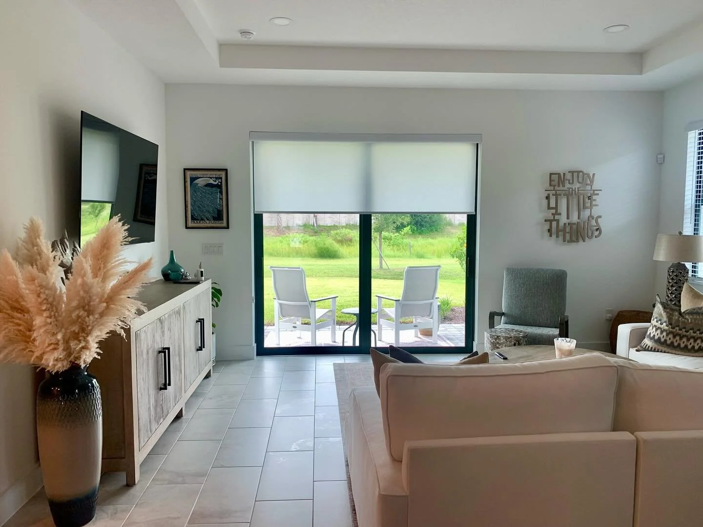 Living room with a sliding glass door with shades ,and a small table, a beige couch, a gray armchair, a media console, a large vase with pampas grass, art on the walls, and a decorative sign that says 'Enjoy the little things'.