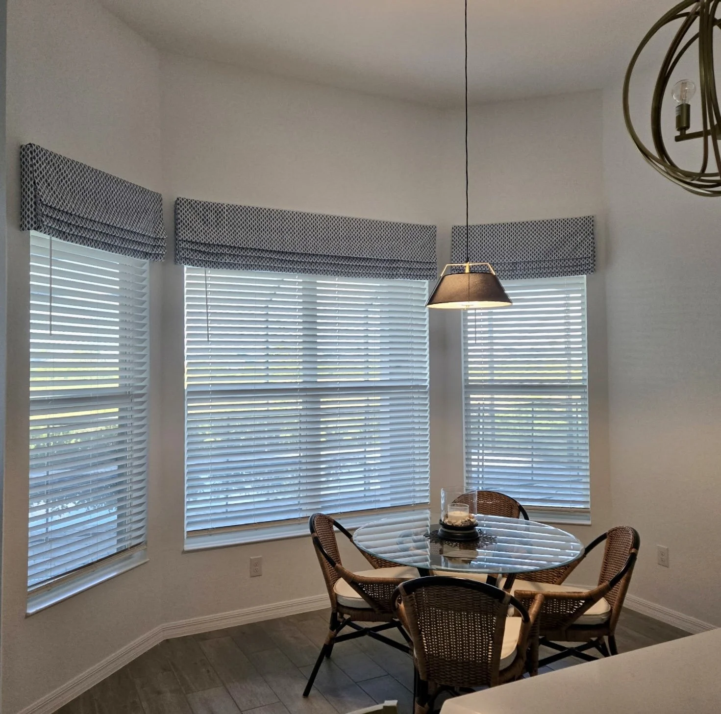 A dining area with a round glass table surrounded by four wicker chairs with white cushions, placed in front of three large windows with white blinds and valances and matching valances, and a hanging light fixture overhead.