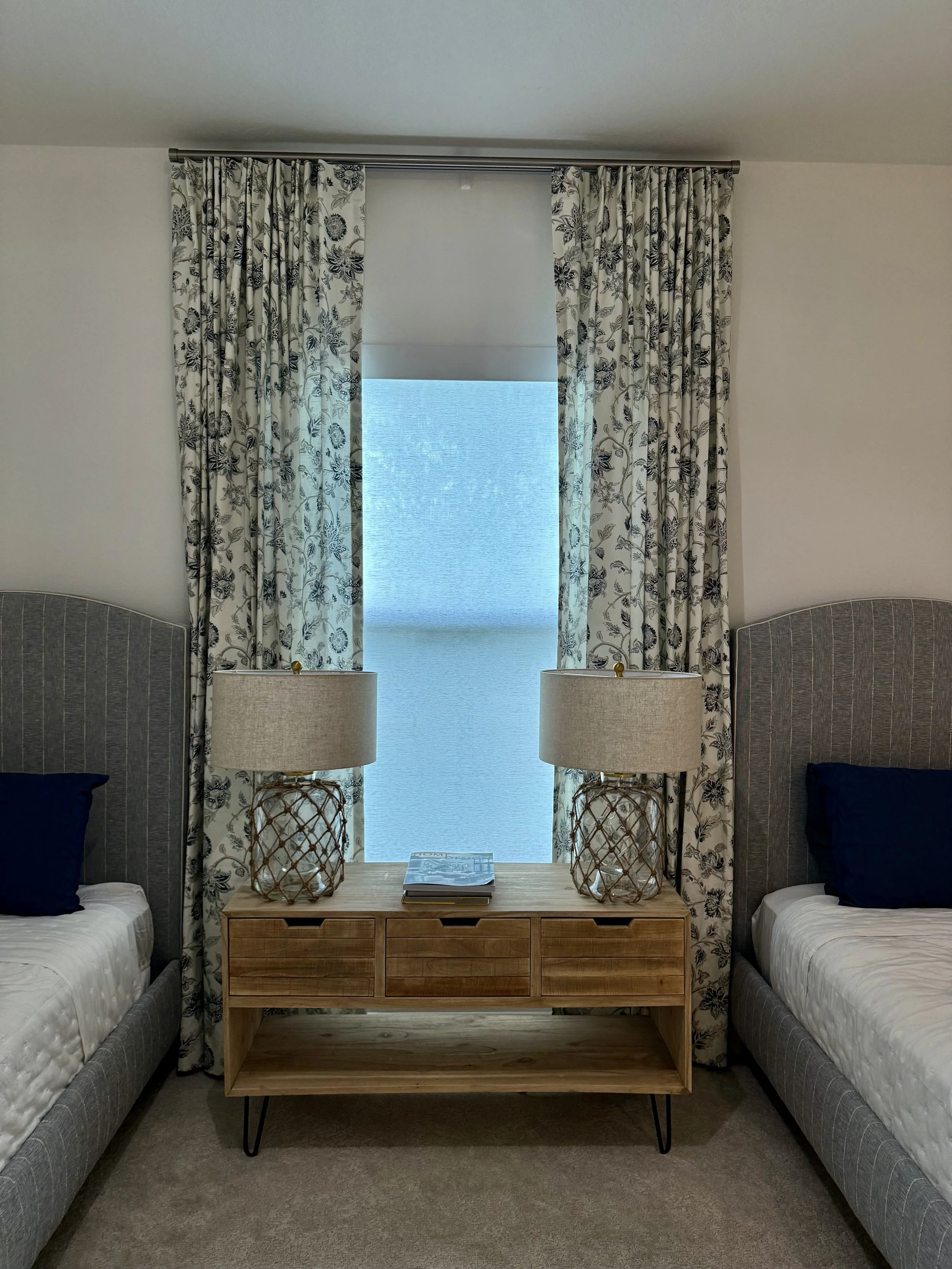 Two twin beds with gray upholstered headboards, navy pillows, a wooden nightstand with two lamps, a window with floral drapes, and a blue roller shade in a bedroom.