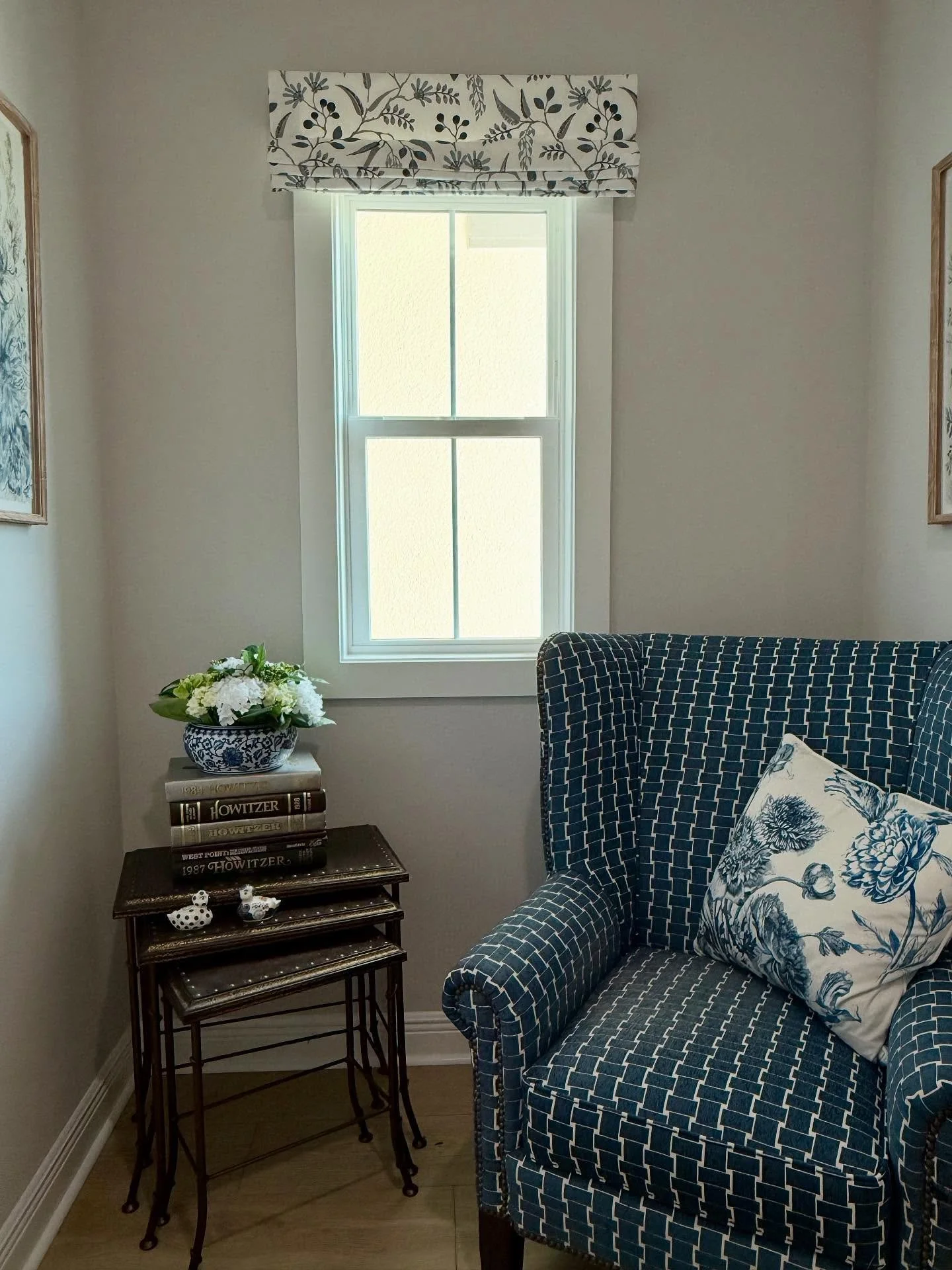 A cozy corner of a room with a window and a seat. The window has a patterned valance, and sunlight is coming through. Next to the window is a small black table with a stack of books, a decorative bowl with flowers, and two small white ceramic birds.