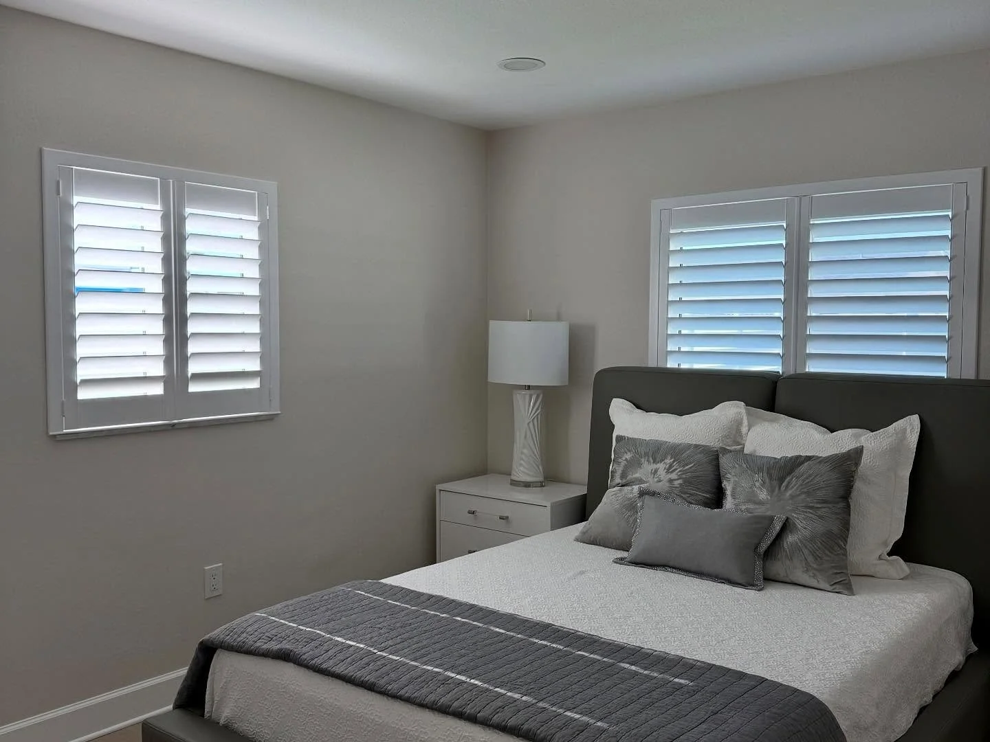 A bedroom with a bed with white and gray bedding, gray pillows, and a gray headboard, a white nightstand with a white lamp, and two windows with white plantation shutters.