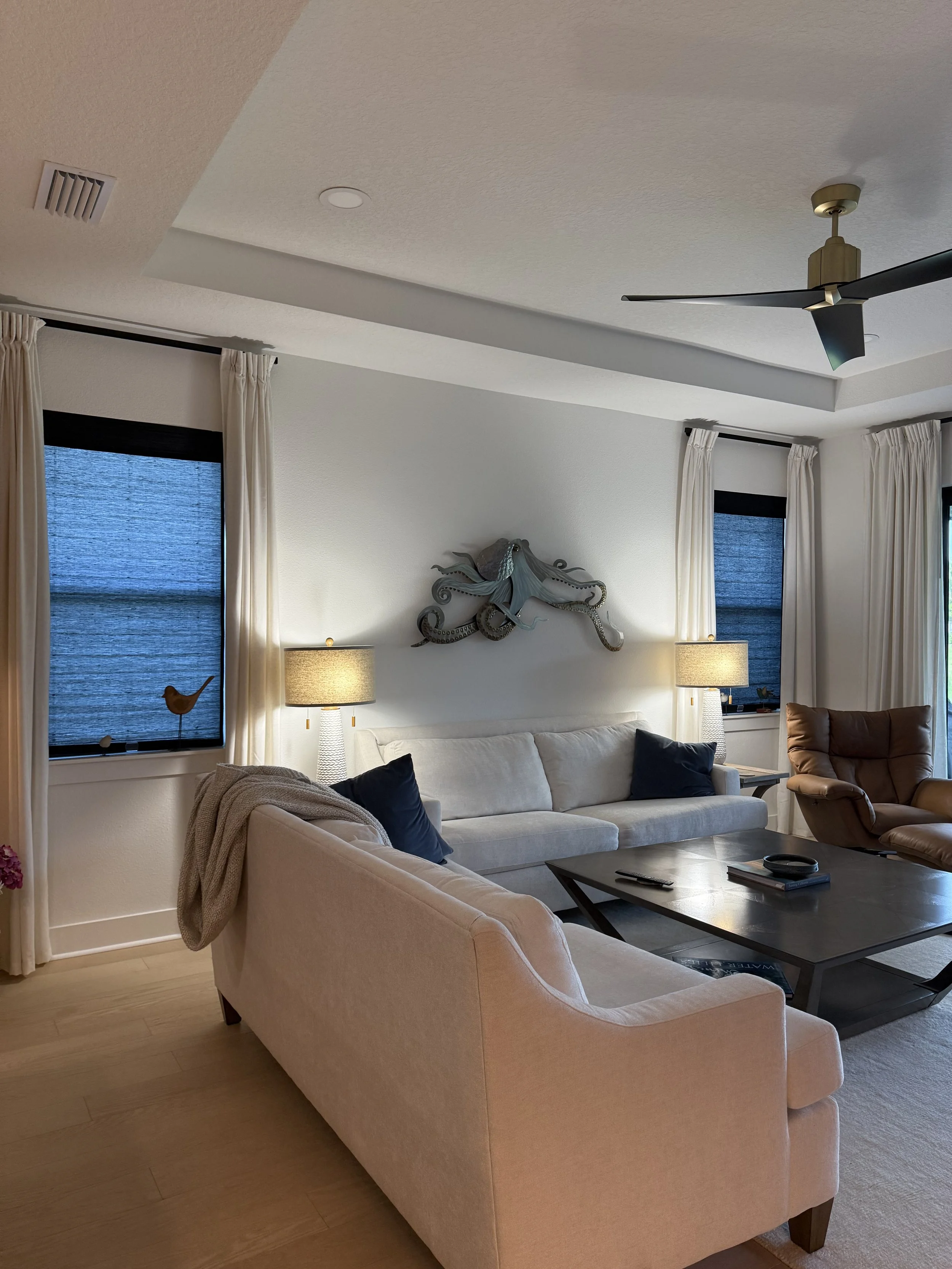 Living room with white sofa, beige armchair, and brown recliner. Two windows with white drapes, lamps on side tables, and a wall-mounted octopus decoration. Wooden coffee table with remote, books, and a decorative dish.