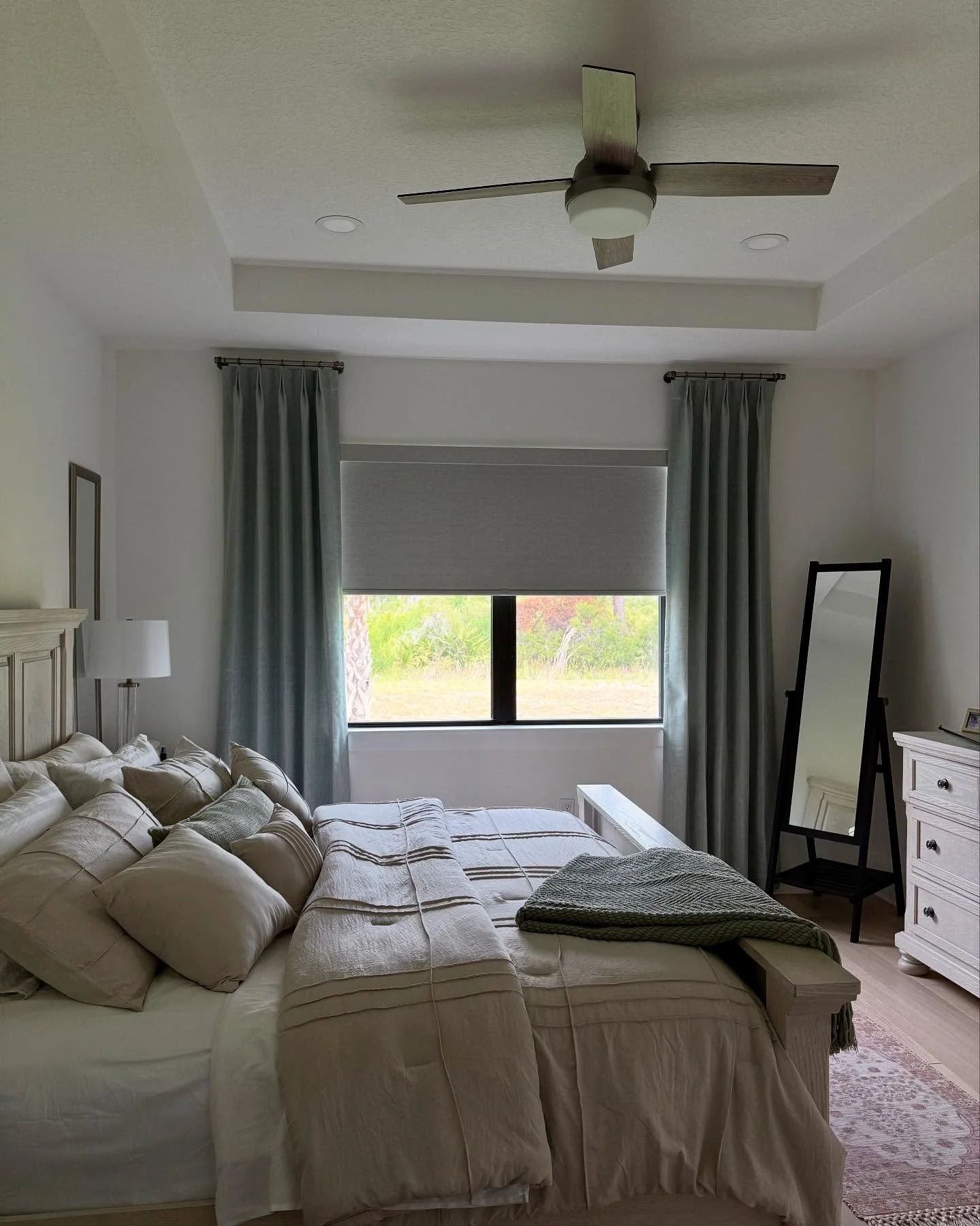 A bedroom with a bed, window with drapes and shades, white dresser, full-length mirror, ceiling fan, and curtains.