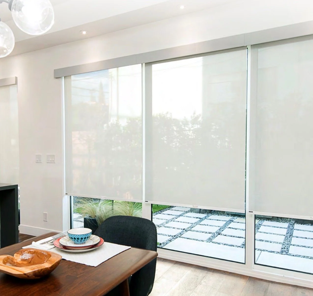 Kitchen or dining area with large sliding glass doors covered by white roller blinds, a wooden dining table set with bowls and plates, and outdoor patio with tiles and greenery visible through the glass.