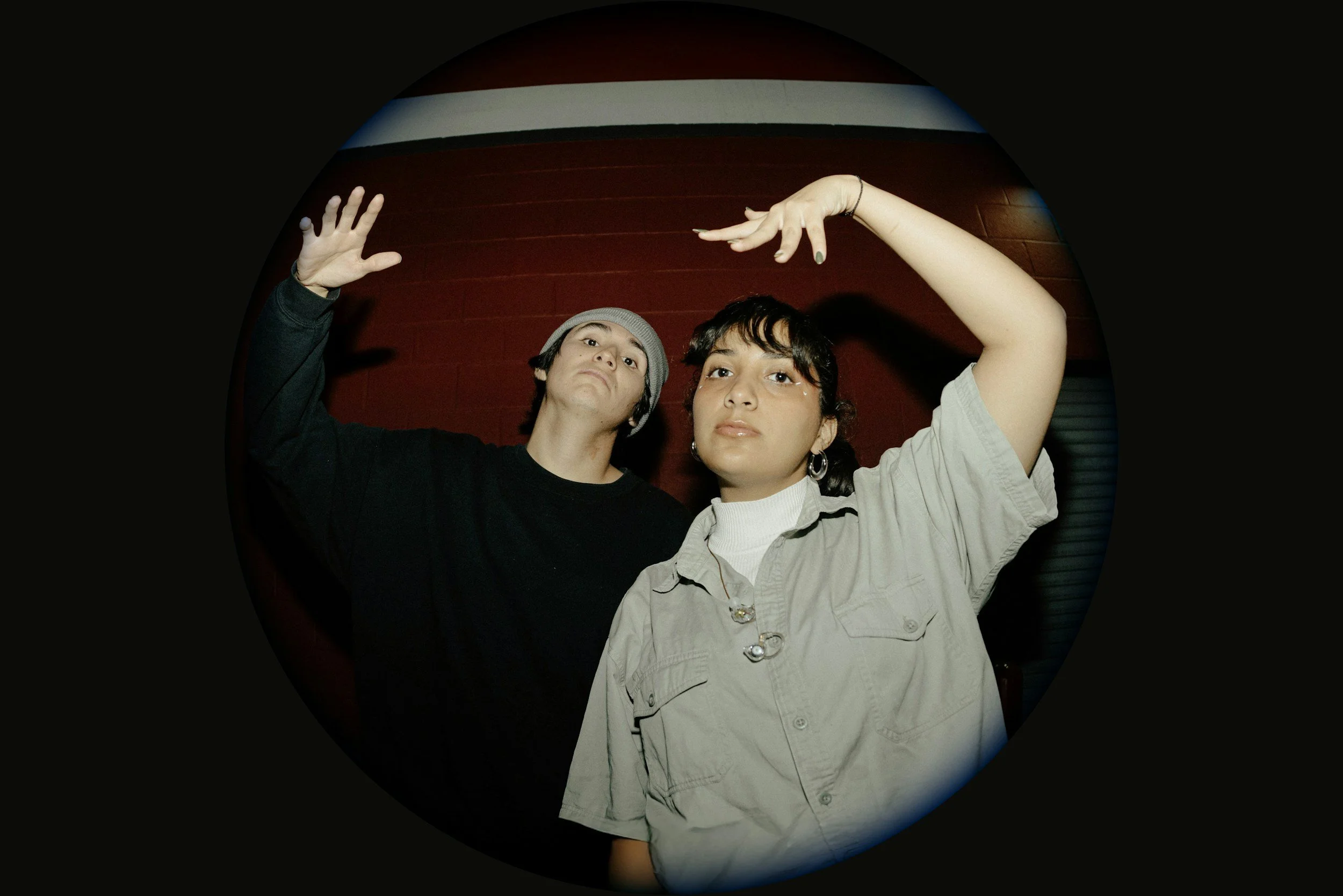 Two young women posing indoors against a red brick wall, one with short dark hair and hoop earrings, wearing a beige shirt, and the other with short dark hair, wearing a beanie and a black shirt, with their hands raised in expressive gestures.