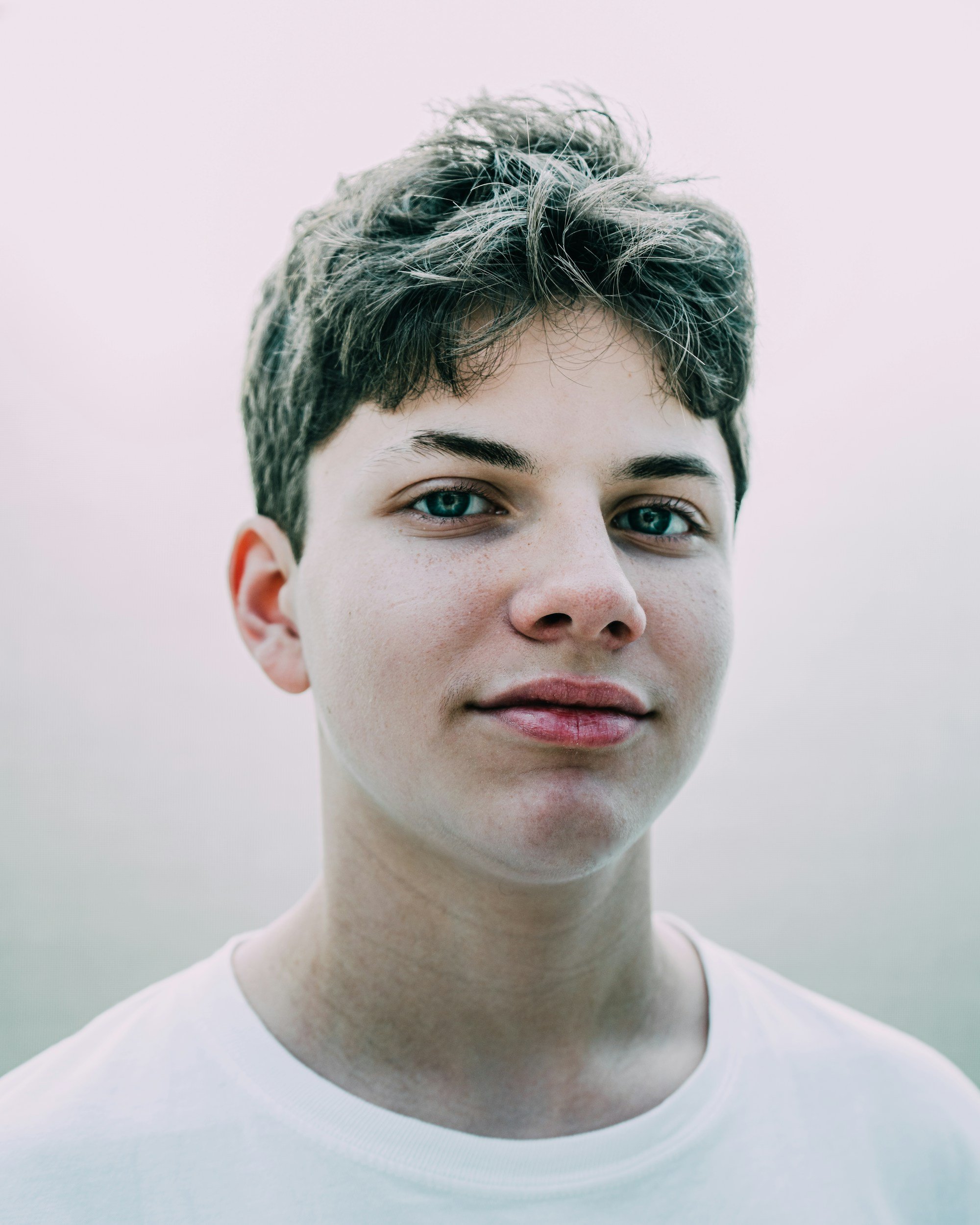 Close-up of a young person with short, wavy brown hair, blue eyes, and a slight smile, wearing a white shirt.