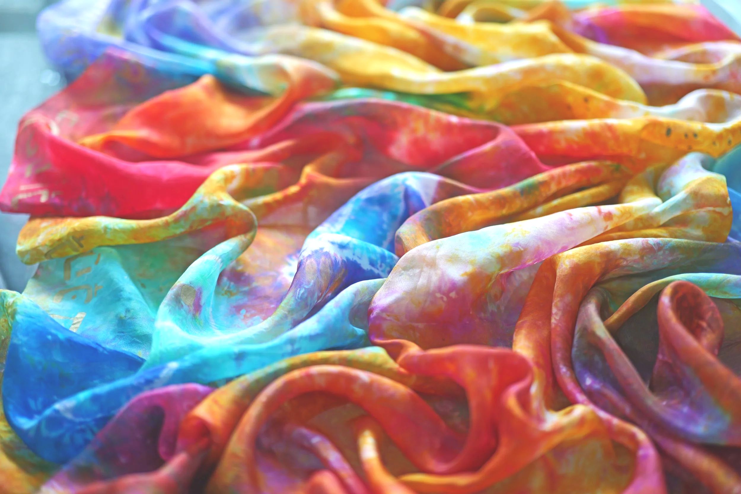 Colorful tie-dye fabric with vibrant swirls of pink, red, orange, yellow, green, blue, and purple, arranged in a loose, textured pattern.
