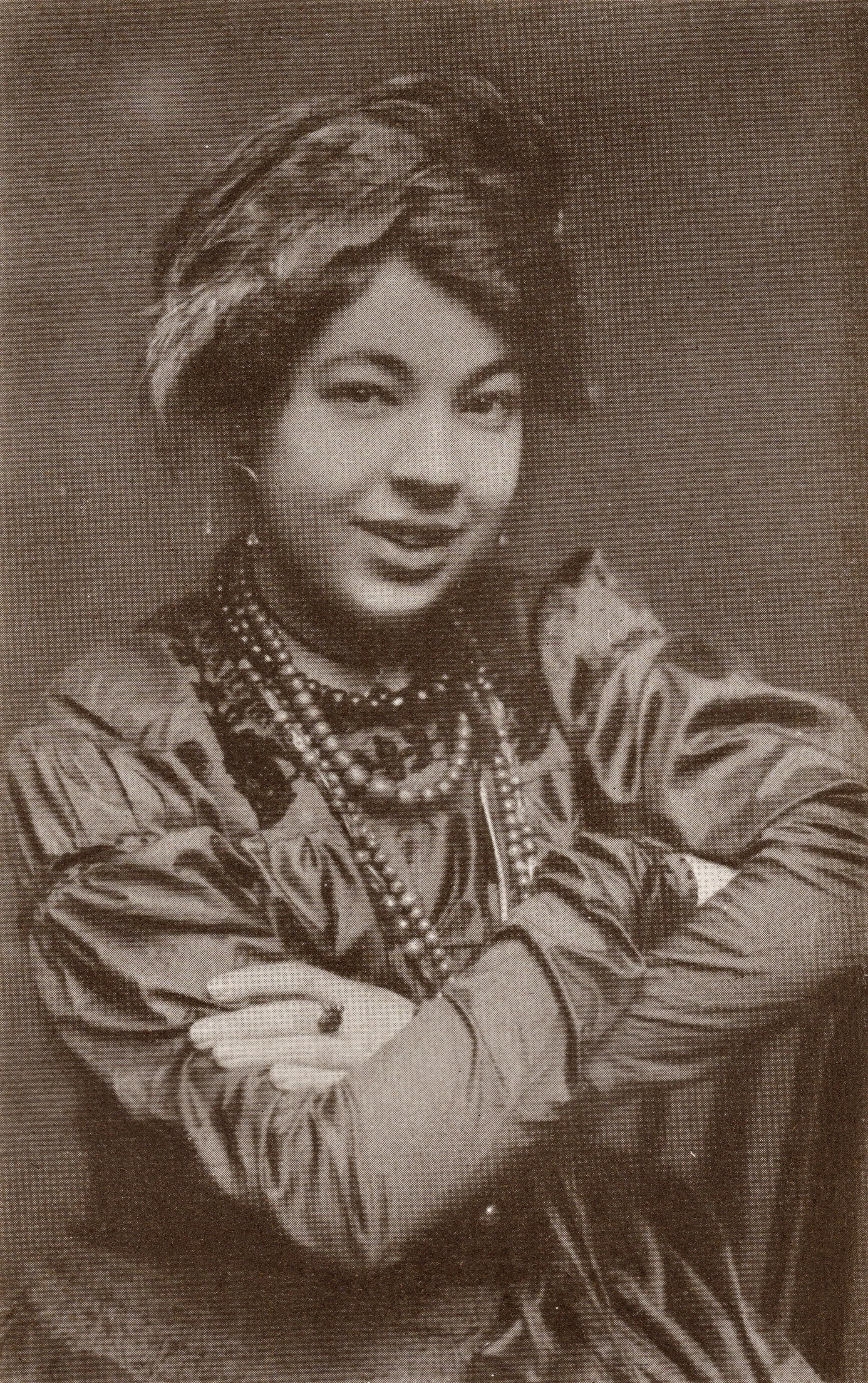 A woman with layered hairstyle, wearing patterned clothing and layered necklaces, smiling with arms crossed.