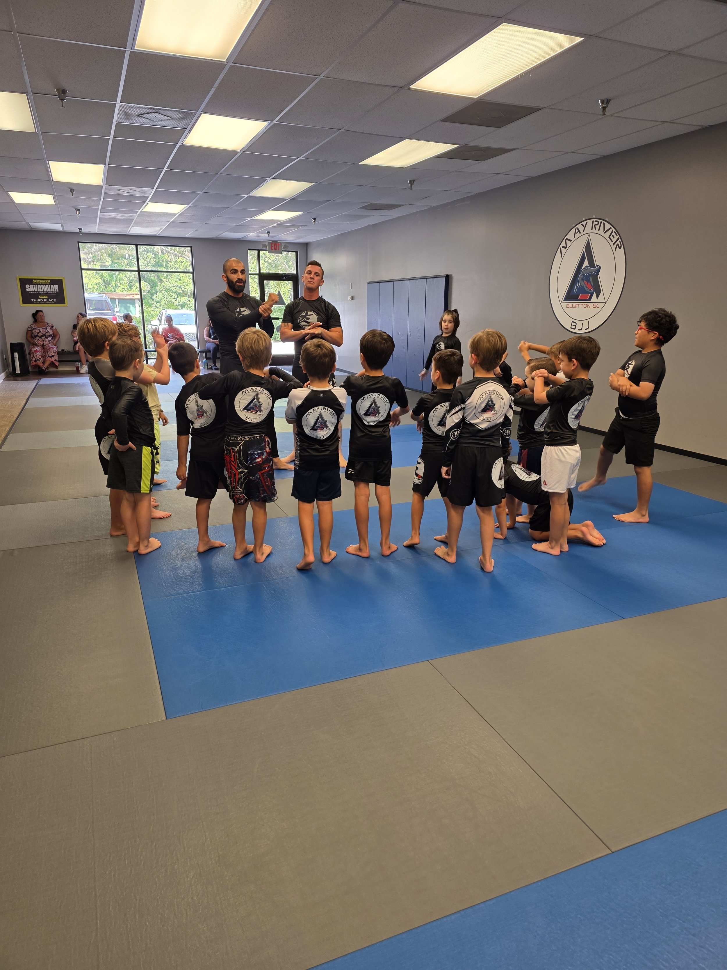 A group of young children in black martial arts uniforms standing on a mat inside a martial arts school, listening to instructors giving a demonstration or instructions.