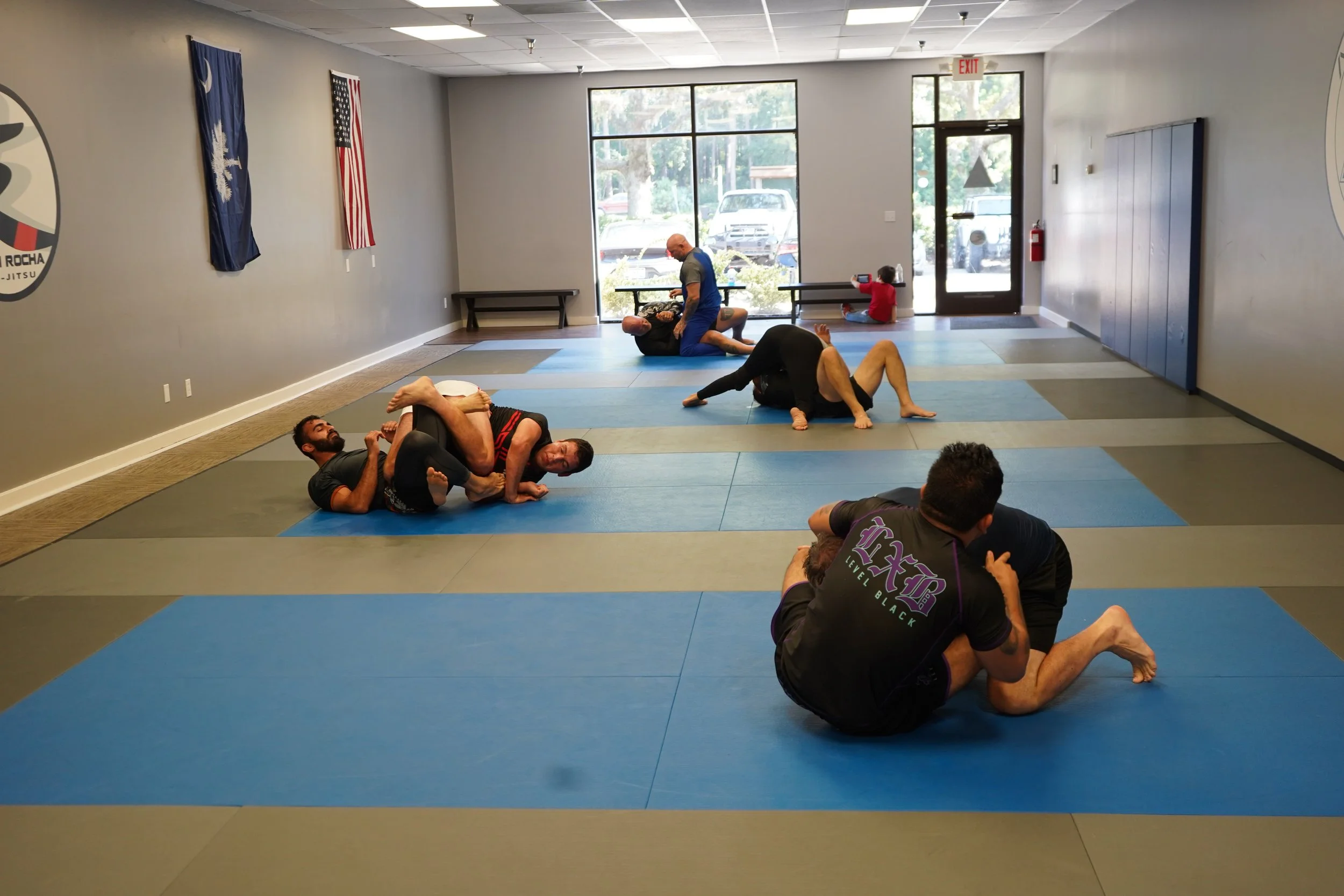 People practicing Brazilian Jiu-Jitsu on padded mats in a martial arts gym during training or class. Some are grappling and some are sitting or lying on the mats.
