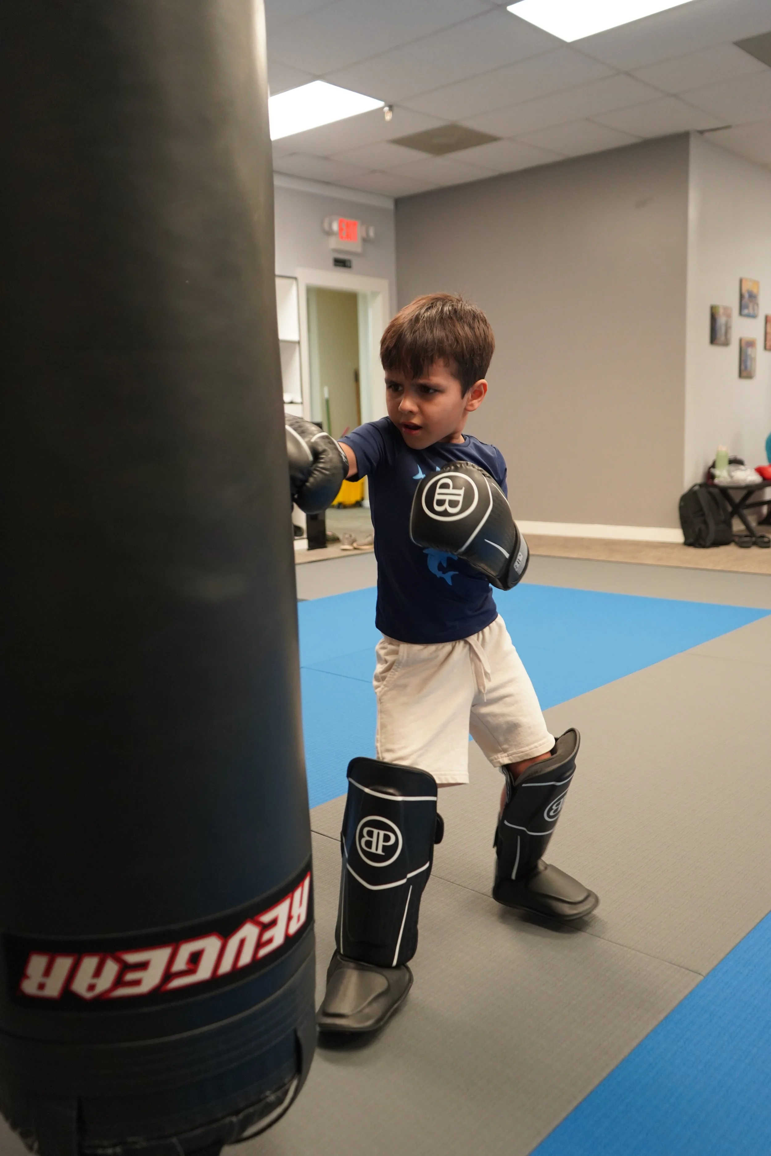 Young boy practicing boxing, wearing boxing gloves and protective gear, punching a punching bag in a gym.